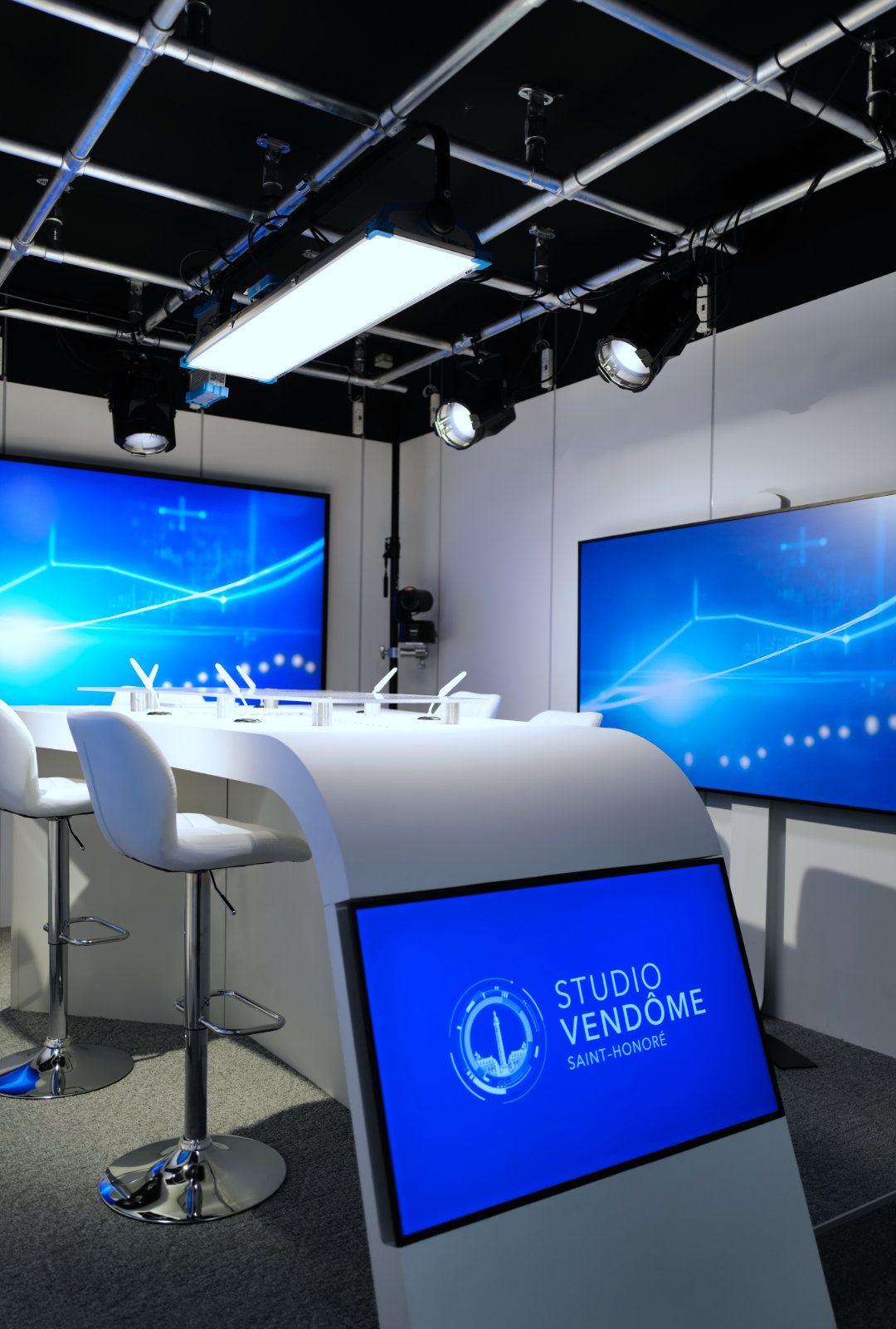 Modern broadcast studio with white curved desk, bar stools, multiple blue digital display screens, and overhead lighting rig.