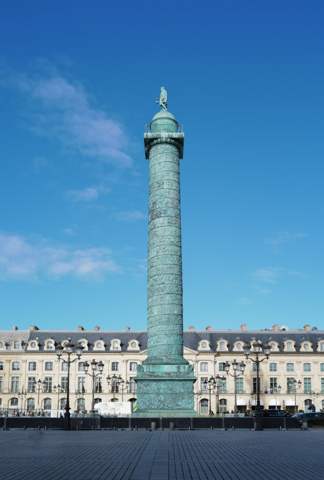 Tall greenish bronze column with detailed carvings and a statue on top, set in a plaza with historic building and blue sky background.