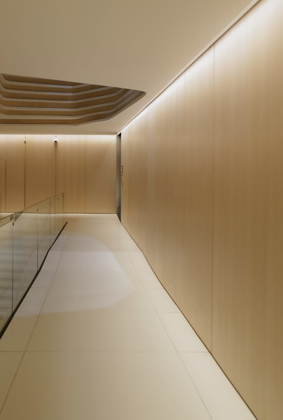 Minimalist hallway with light wooden walls, white tiled floor, glass railing on the left, and recessed ceiling design.