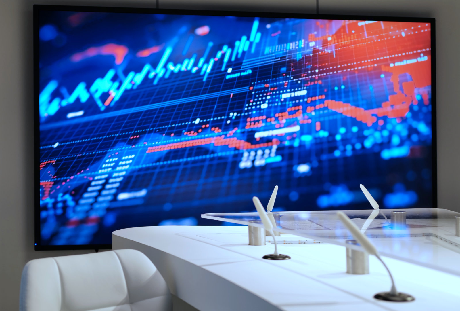 Modern office desk with white chairs and adjustable microphones in front of a large digital screen displaying blue and red financial graphs and data.