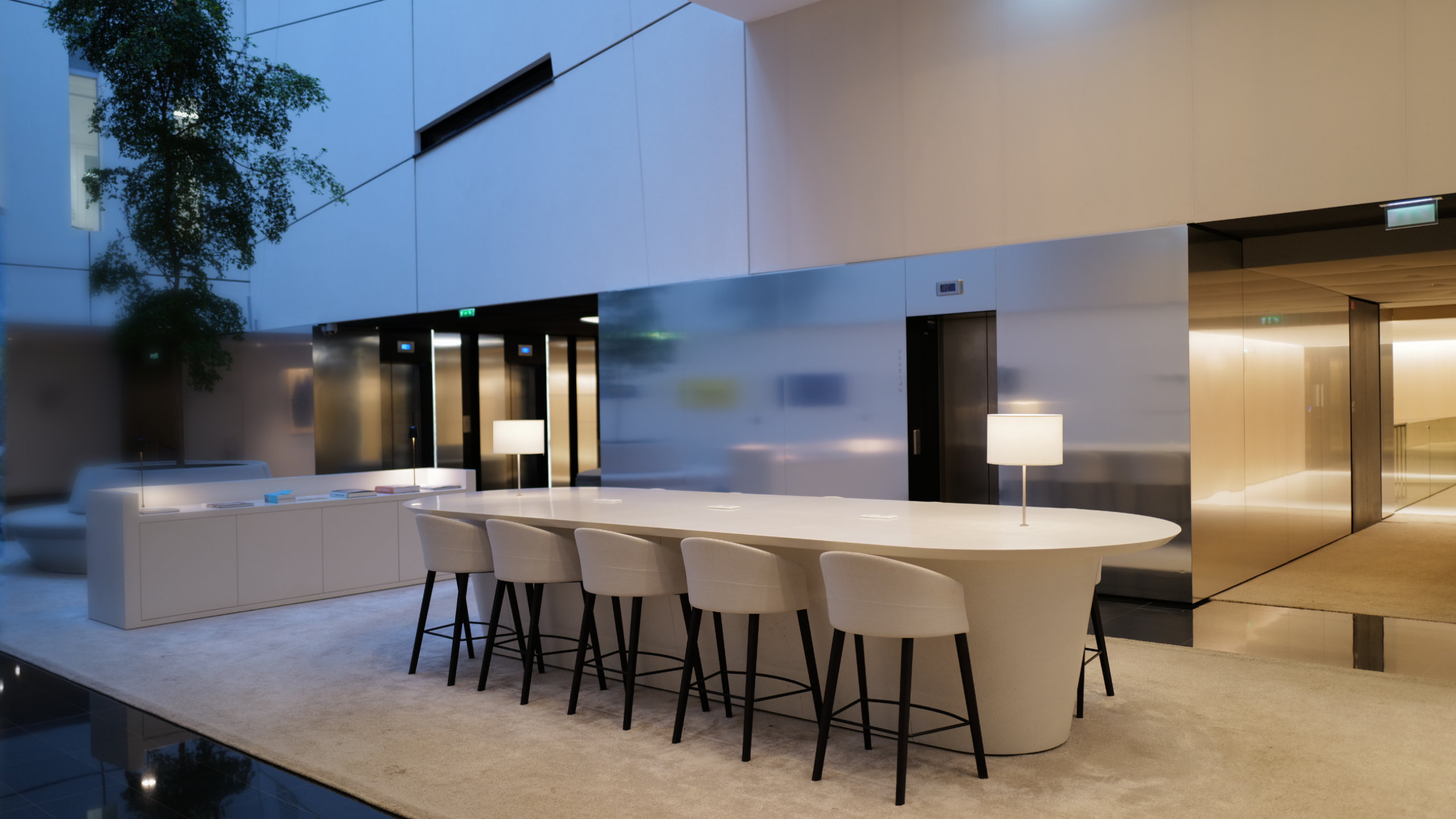 Modern office lobby with a long white table, six cushioned chairs, two table lamps, and a tree in the corner.