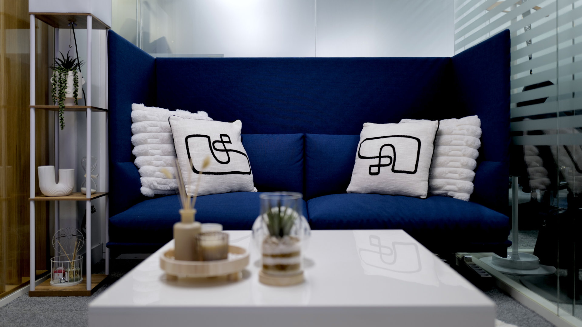 Modern office lounge with a blue sofa adorned with white textured pillows, a white table with decorative items, and a wooden shelf with plants.