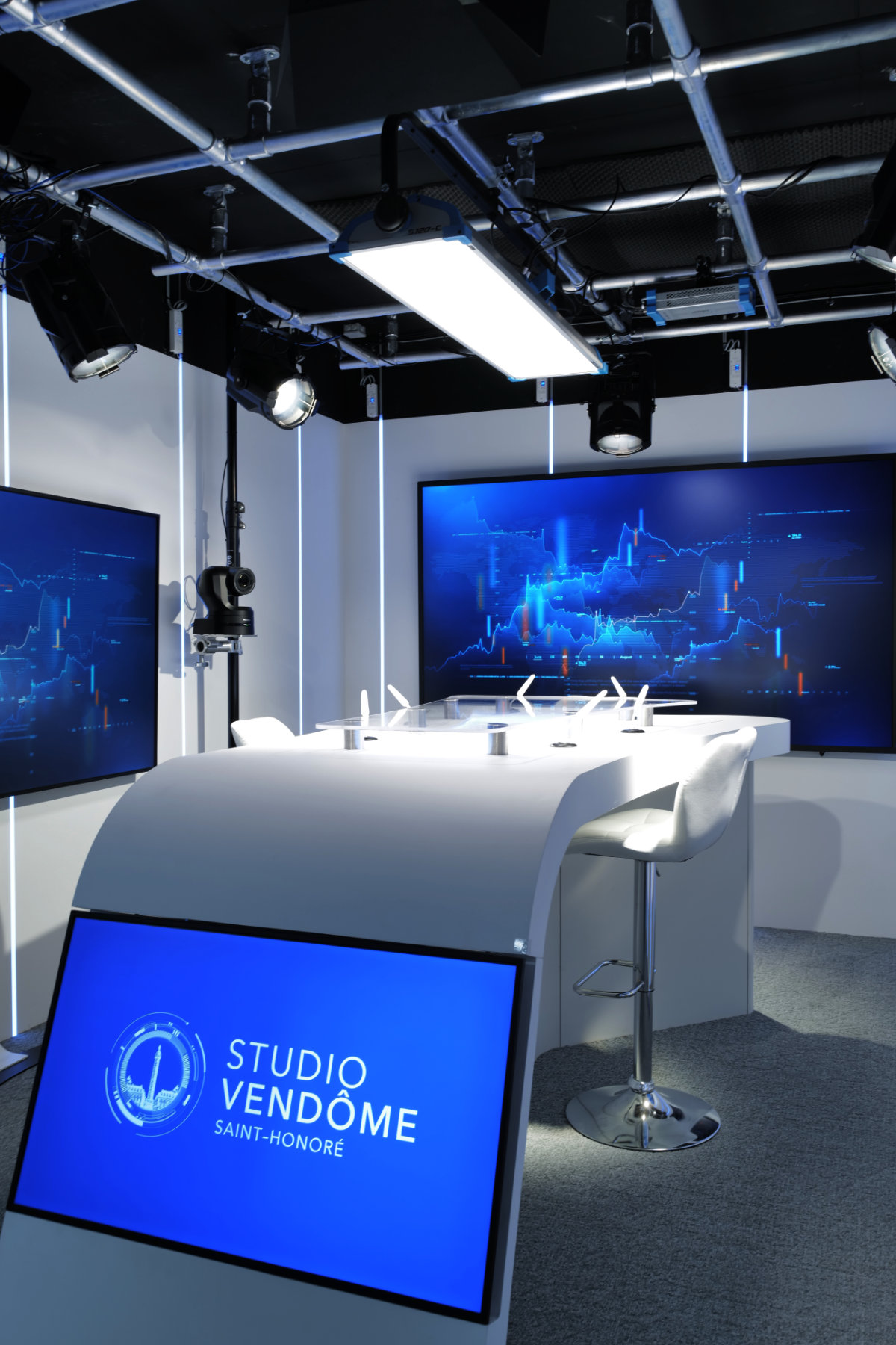 'Studio Vendôme Saint-Honoré' setup with curved white desk, multiple large screens displaying blue data charts, and a chair on a gray carpet.