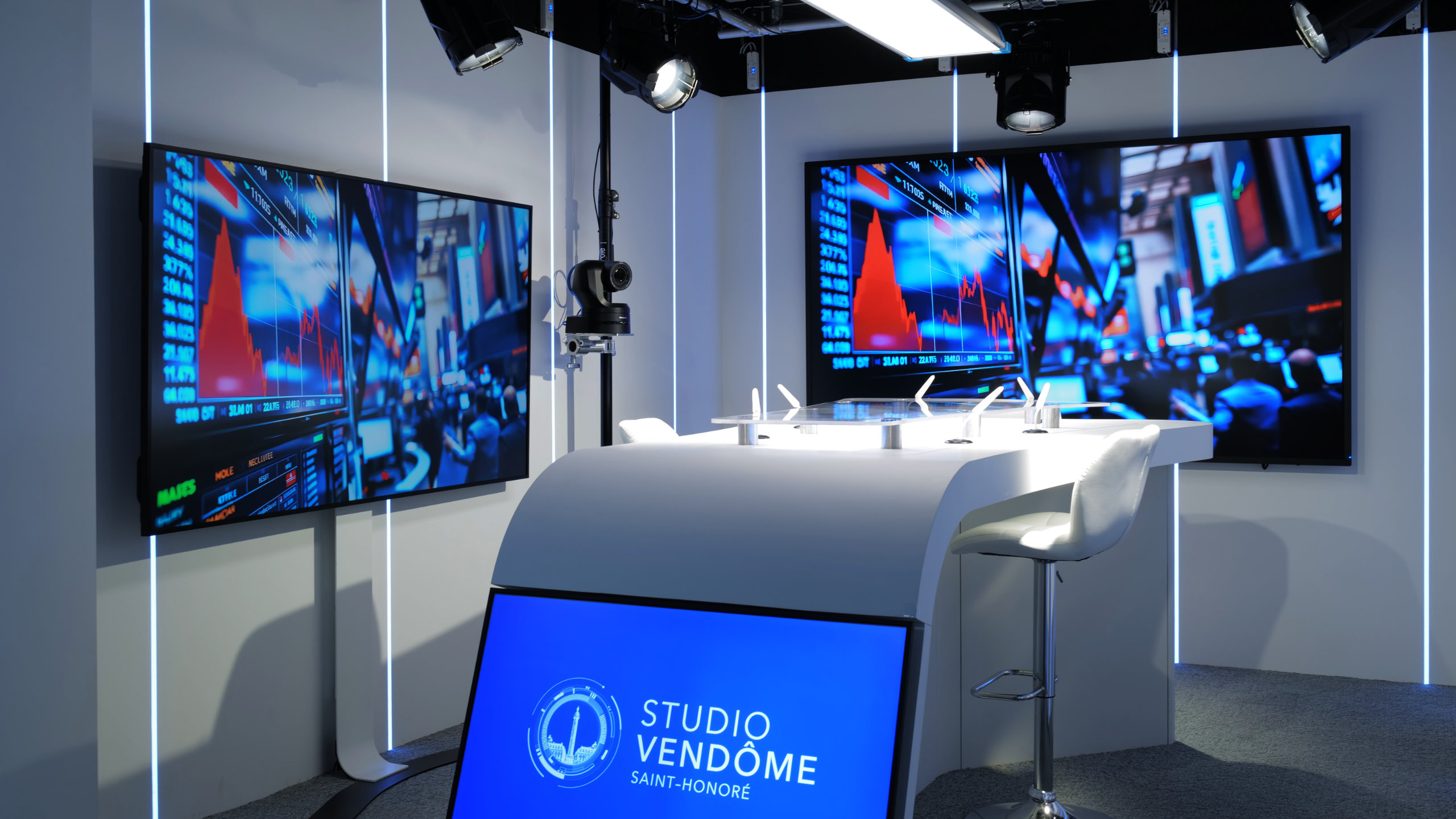 'Studio Vendôme Saint-Honoré' Modern studio set with white curved desk, two large screens displaying financial graphs, and a chair.