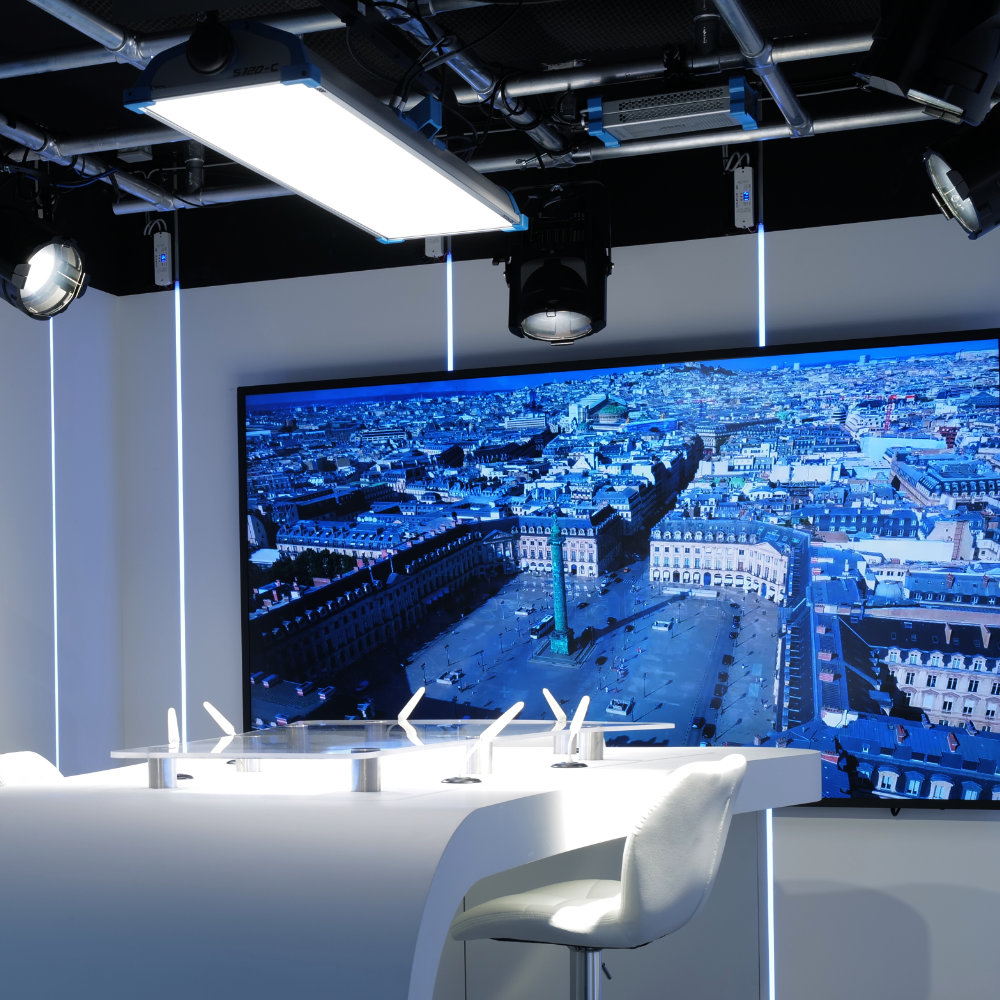Modern studio  'Studio Vendôme Saint-Honoré' setup with a curved white desk, a single white chair, ceiling lights, and a large screen displaying an aerial cityscape.