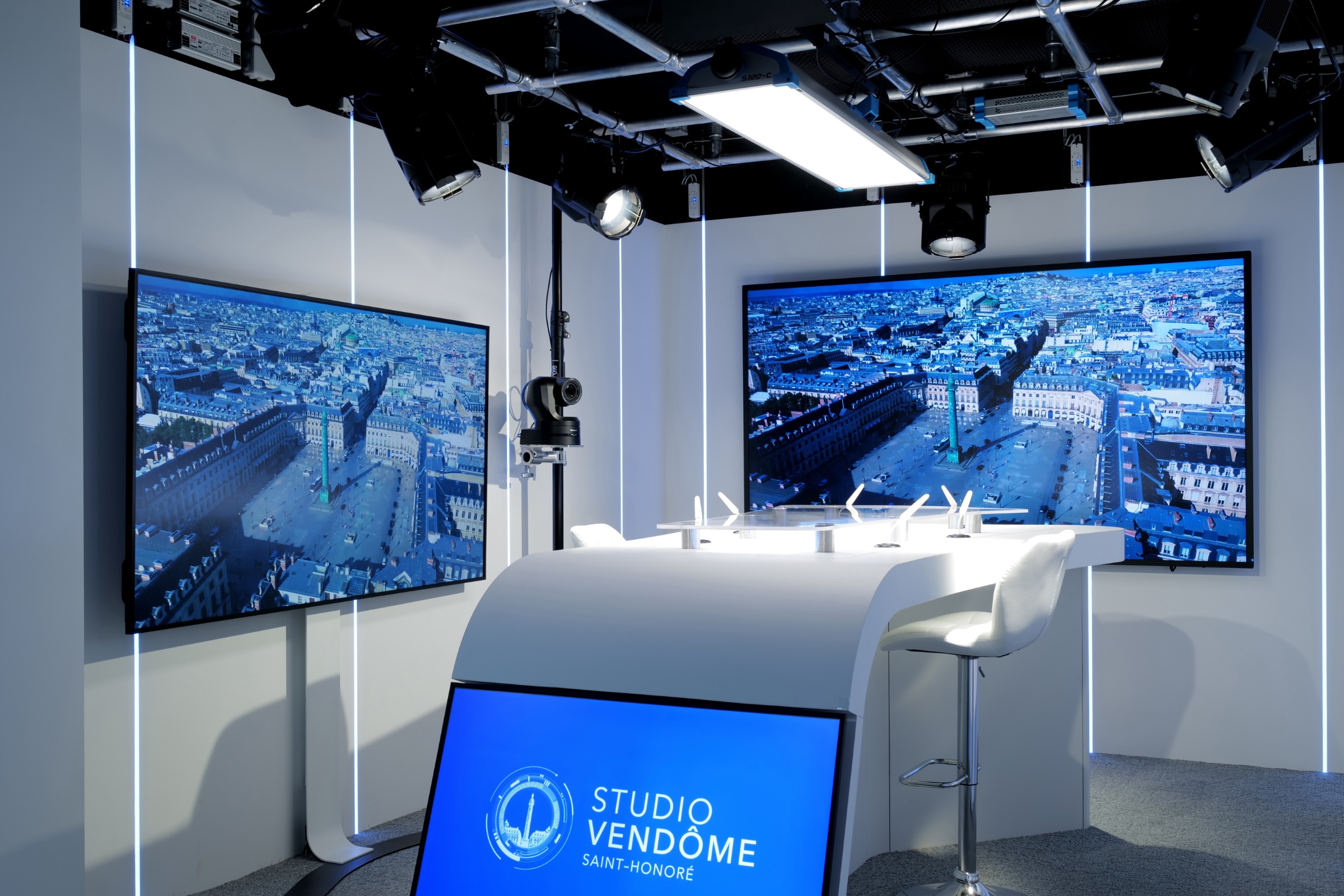 Studio TV professionnel
Vendôme Saint-Honoré set with a curved white desk, a white chair, and multiple large screens showing aerial views of an urban square.