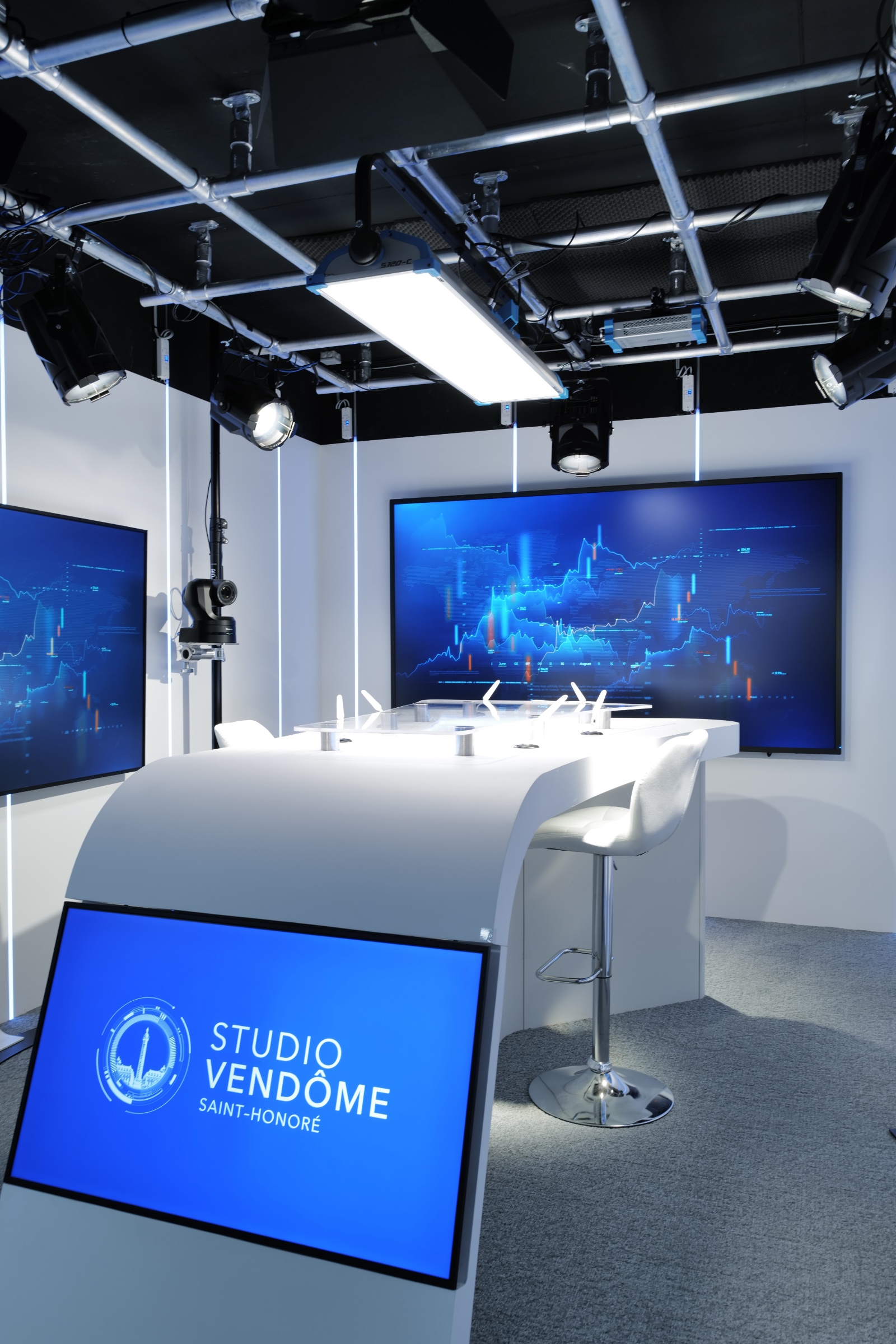 Modern broadcast studio with curved white desk, multiple screens showing blue financial charts, studio lighting, and a sign reading 'Studio Vendôme Saint-Honoré'.