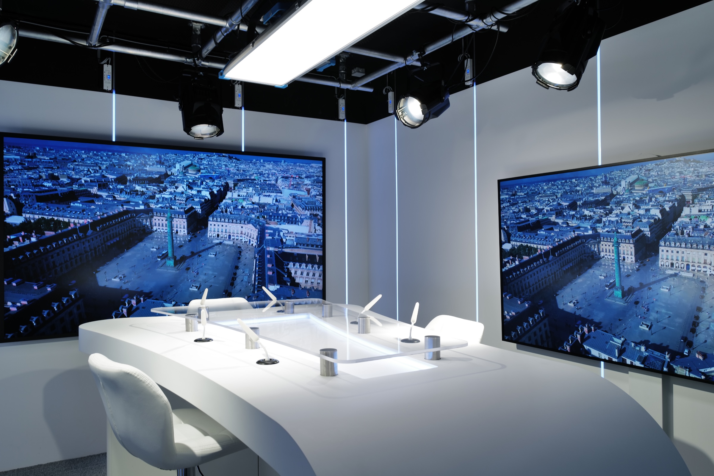 Studio TV professionnel
Vendôme Saint-Honoré - Modern white broadcast or podcast studio with two large monitors displaying aerial views of a city square and multiple microphones on a curved white desk.