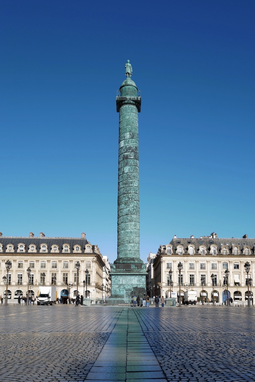 Place Vendôme - Tall bronze column topped with a statue in a large open square flanked by classical buildings under a clear blue sky.