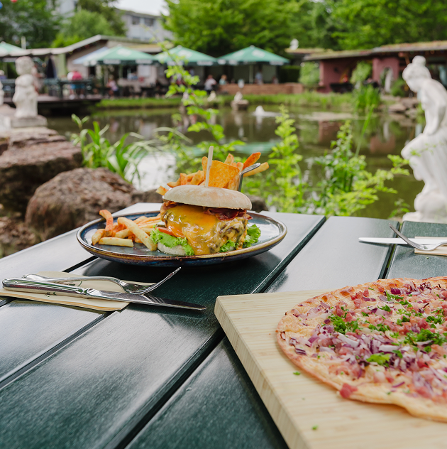 Ein Flammkuchen Elsässer Art auf einem Holzbrett und ein hausgemachter Cheeseburger mit Pommes frites auf einem blauen Teller stehen auf einem Holztisch. Im Hintergrund ein großer Teich und Sonnenschirme
