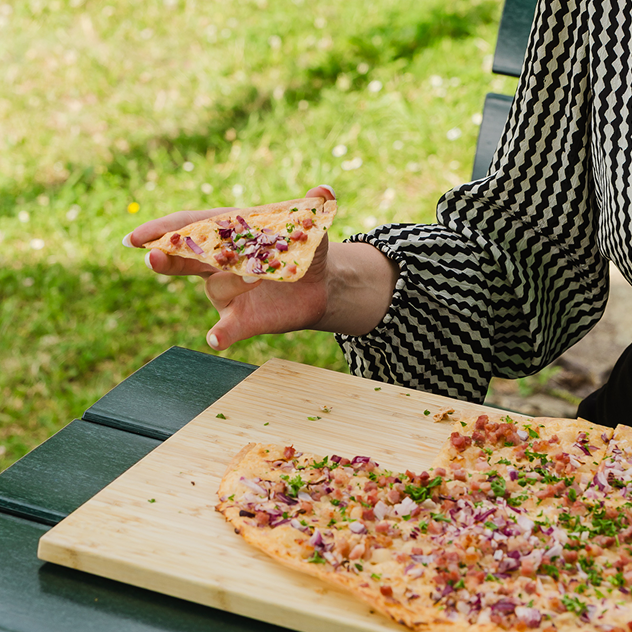 Eine Frau isst einen frischen Flammkuchen Elsässer Art von einem Holzbrett im Biergarten CONTEL. Im Hintergrund grüne Wiese.