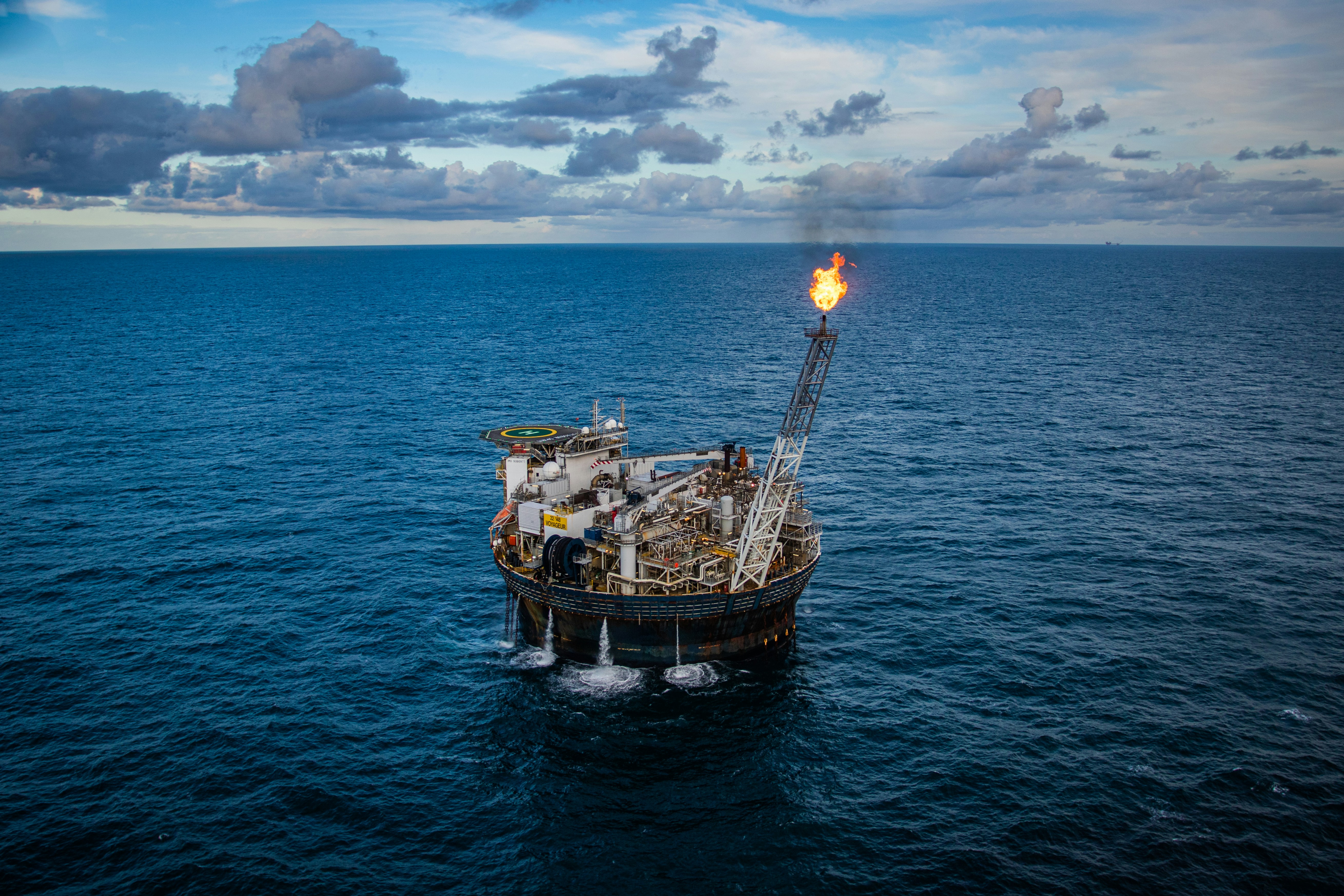 Offshore oil platform floating on calm ocean with a flame burning on a tall flare stack under a cloudy sky.