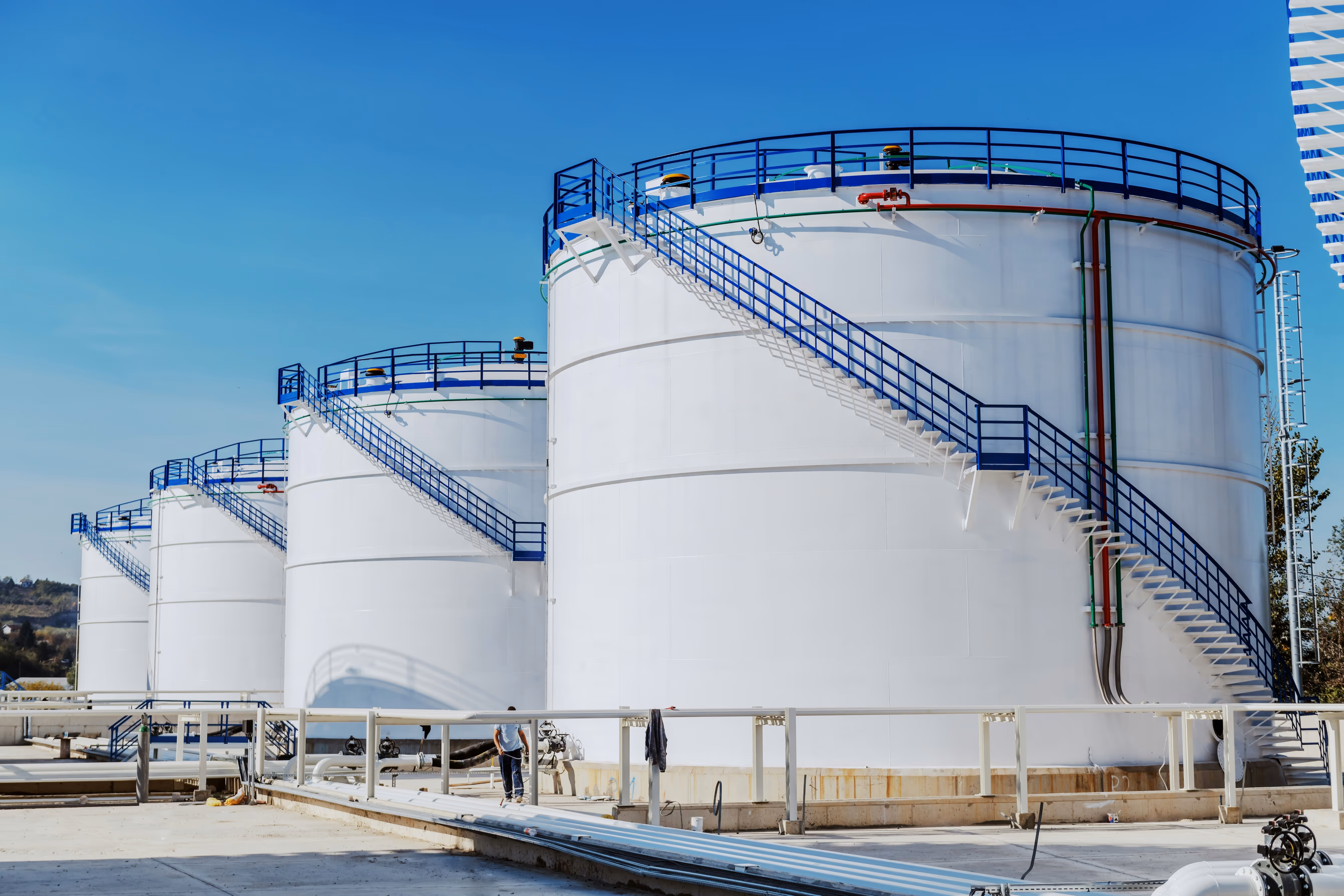 Four large white industrial storage tanks with blue railings and staircases under a clear blue sky.