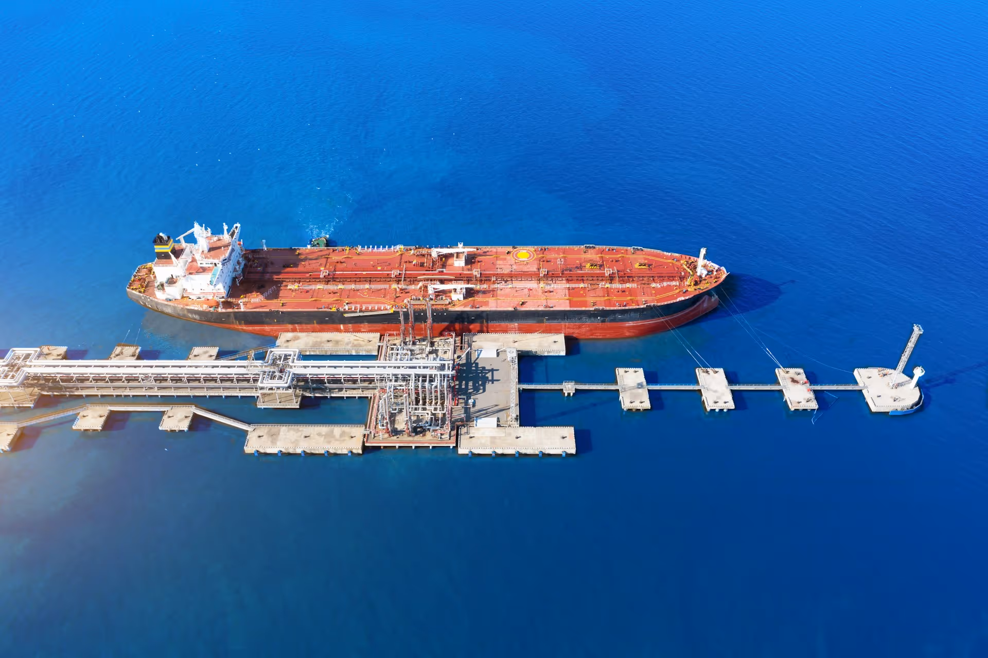 Aerial view of a large red and black tanker ship docked at an industrial pier in deep blue ocean water.