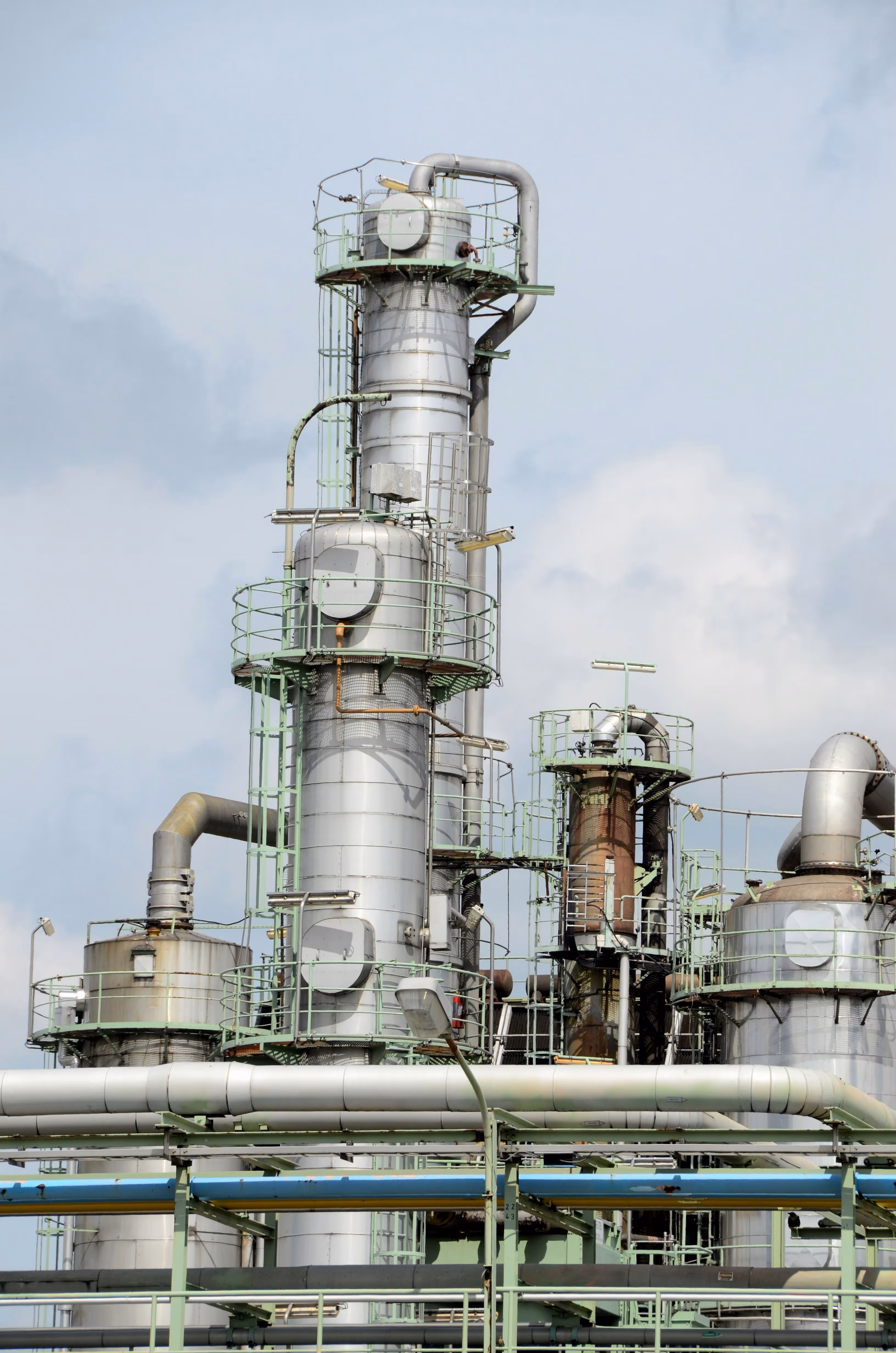 Industrial facility with tall metal distillation towers and interconnected pipes under a cloudy sky.