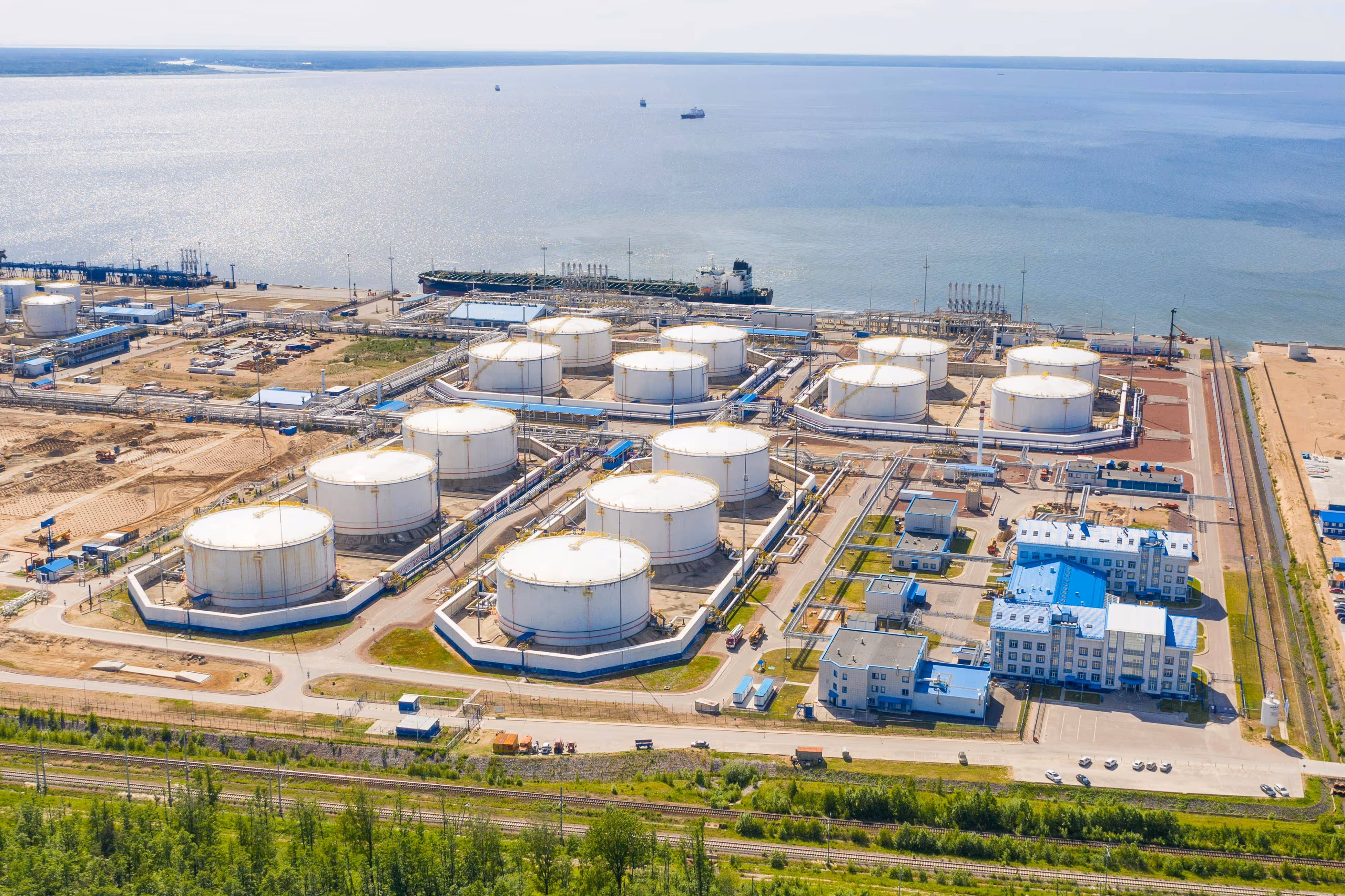 Aerial view of a coastal industrial oil refinery complex with large white storage tanks, buildings with blue roofs, and a ship docked by the water.