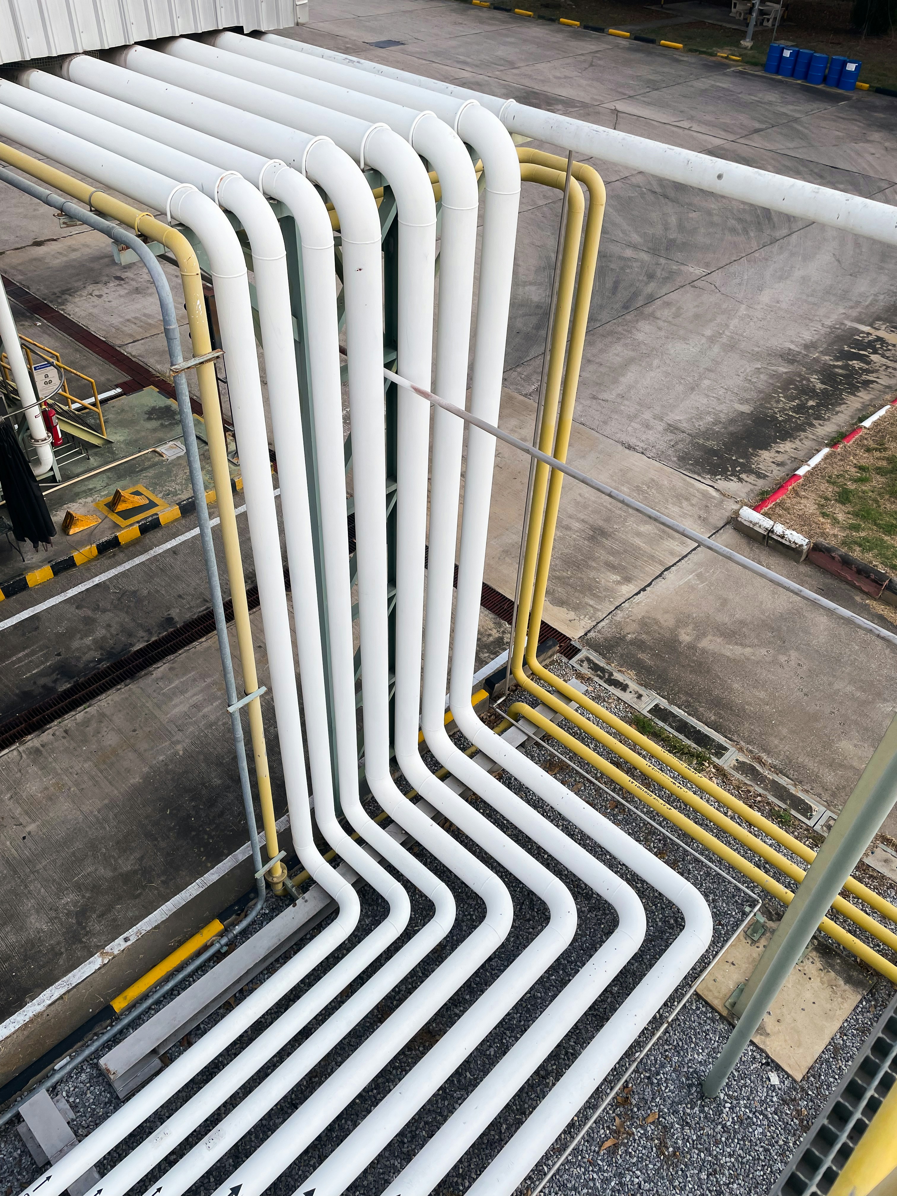Set of large industrial white and yellow pipelines with 90-degree bends arranged in parallel on an outdoor concrete and gravel surface.