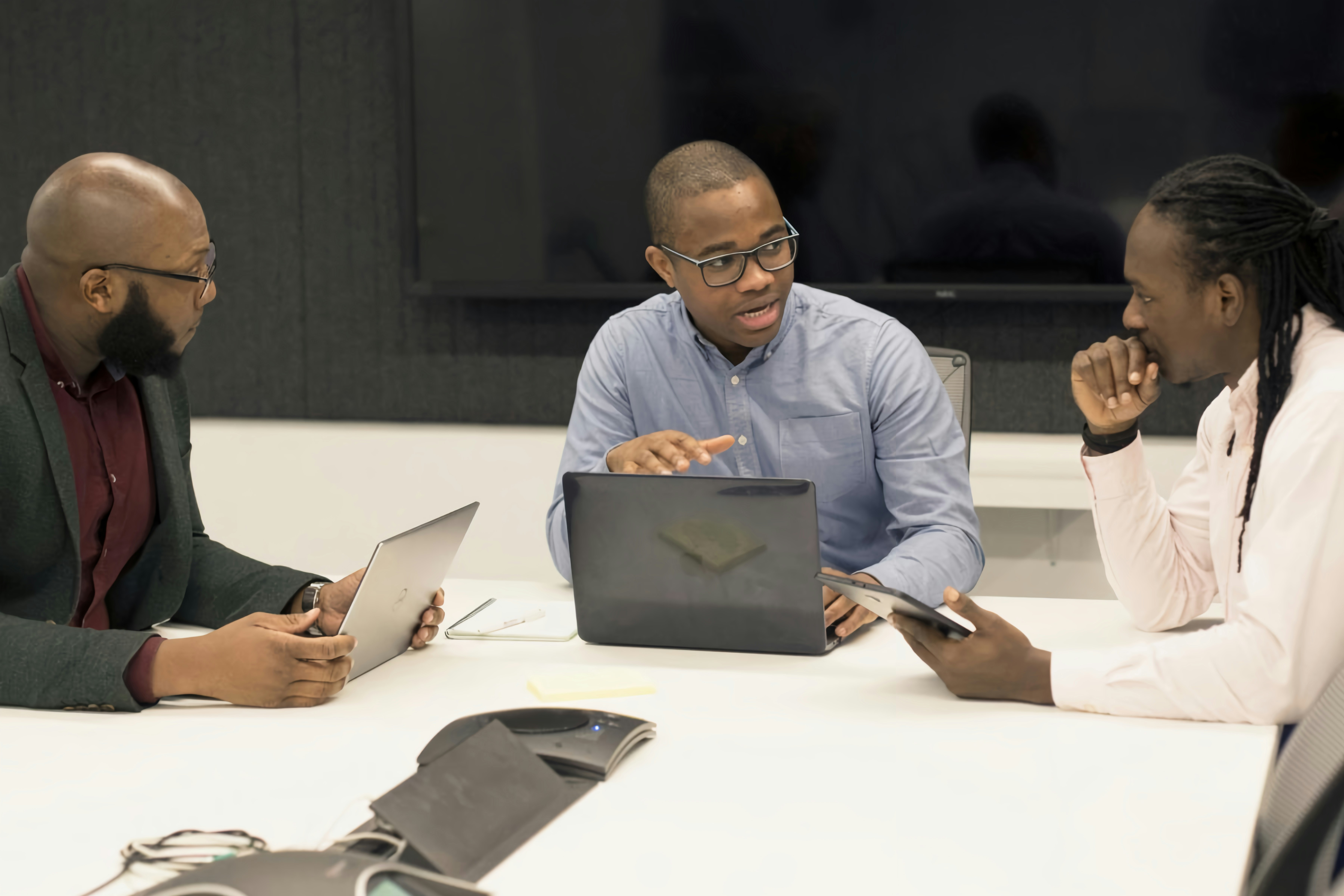 Three men in a meeting room engaged in discussion, using laptops and a tablet.