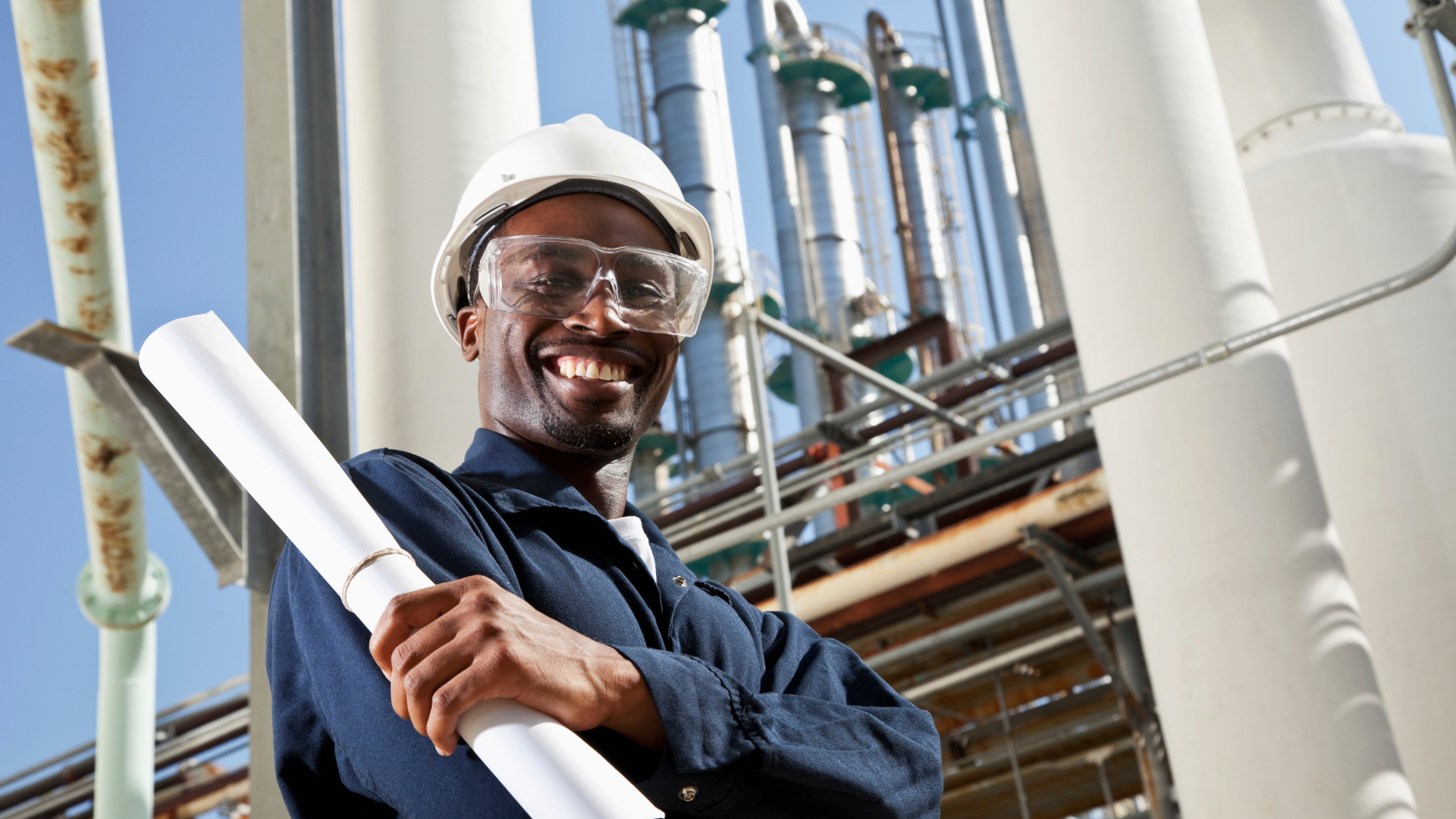 Smiling refinery worker wearing a white hard hat and safety glasses holding rolled blueprints at an industrial site.