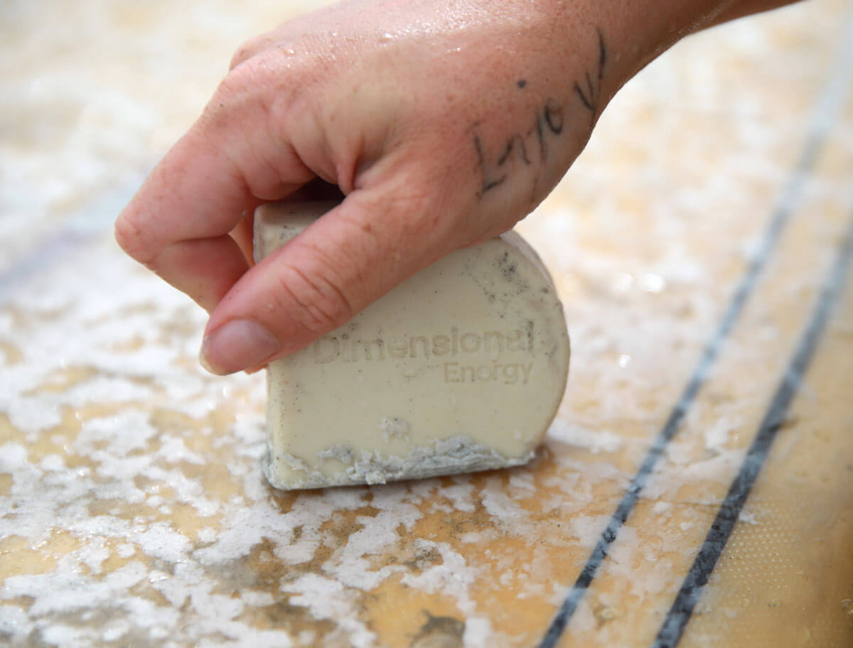 A woman applies Dimensional Energy Surf Wax to her surfboard.