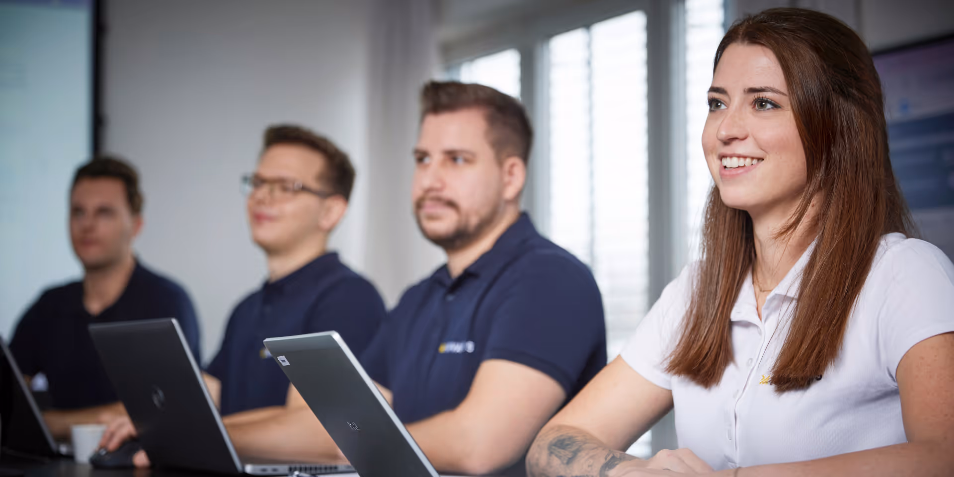 Vier Personen bei einem Meeting, drei Männer in blauen Poloshirts und eine lächelnde Frau in weißem Poloshirt mit Tätowierung am Arm.