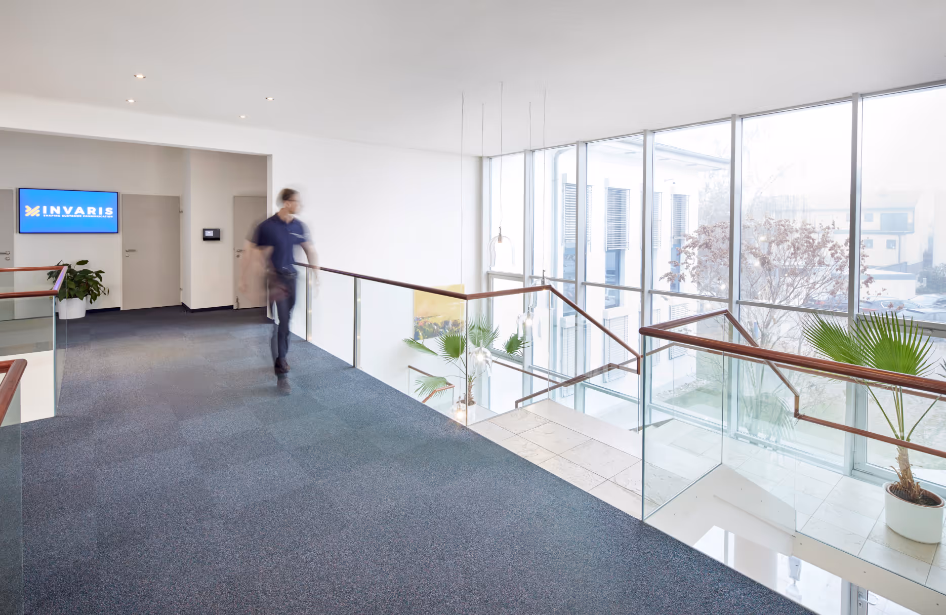 Heller, moderner Büroflur mit blauer Teppichboden, Glasgeländer und großen Fenstern, ein unscharfer Mann geht entlang des Flurs.