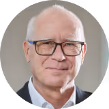 Franz Guszich mit Brille und dunklem Sakko vor unscharfem Hintergrund.