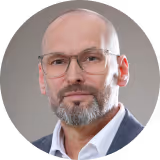 Porträt von Andreas Zsoldos mit Brille in weißem Hemd und dunklem Sakko vor grauem Hintergrund.