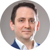 Porträt von Raphael Stracke mit dunklem Haar, weißem Hemd und blauem Sakko vor hellem Hintergrund.