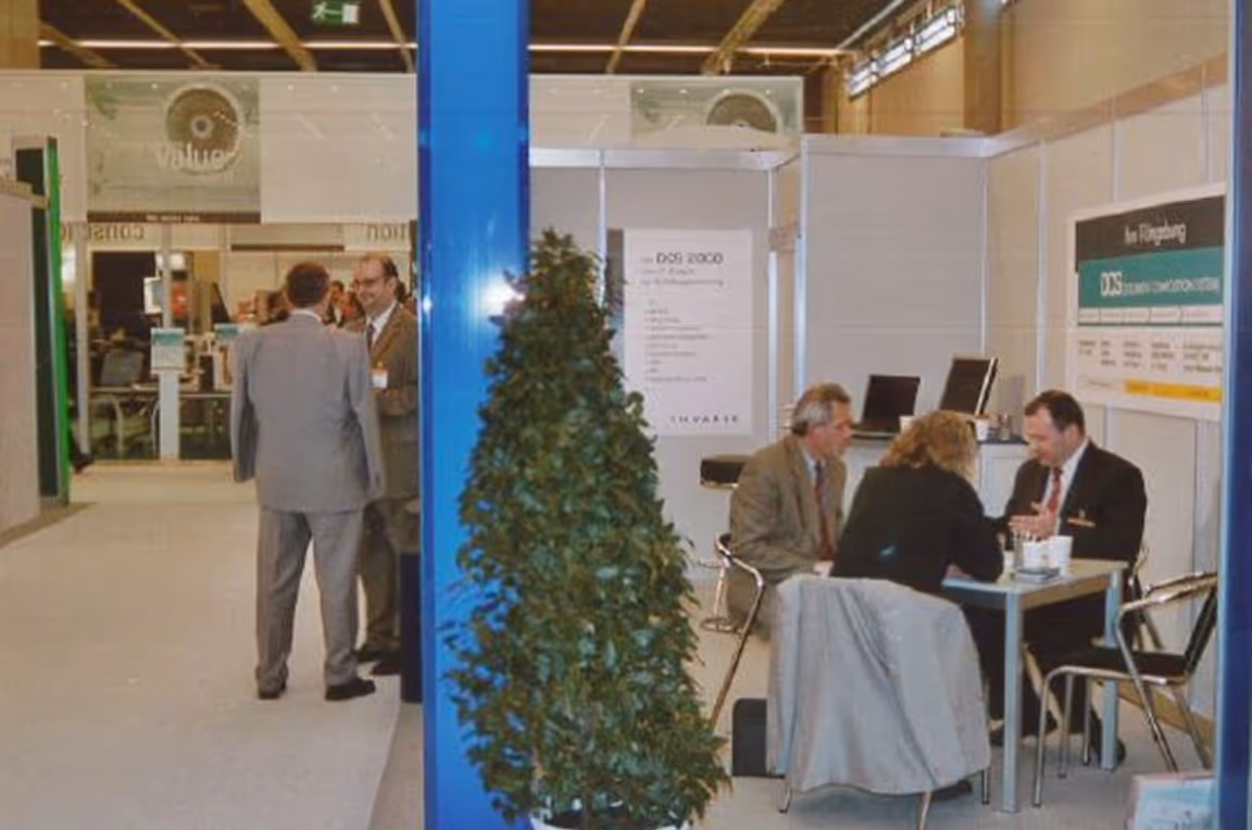 Menschen in Anzügen führen Gespräche in einer Messestandumgebung mit weißen Stellwänden und einem Tisch mit Stühlen.