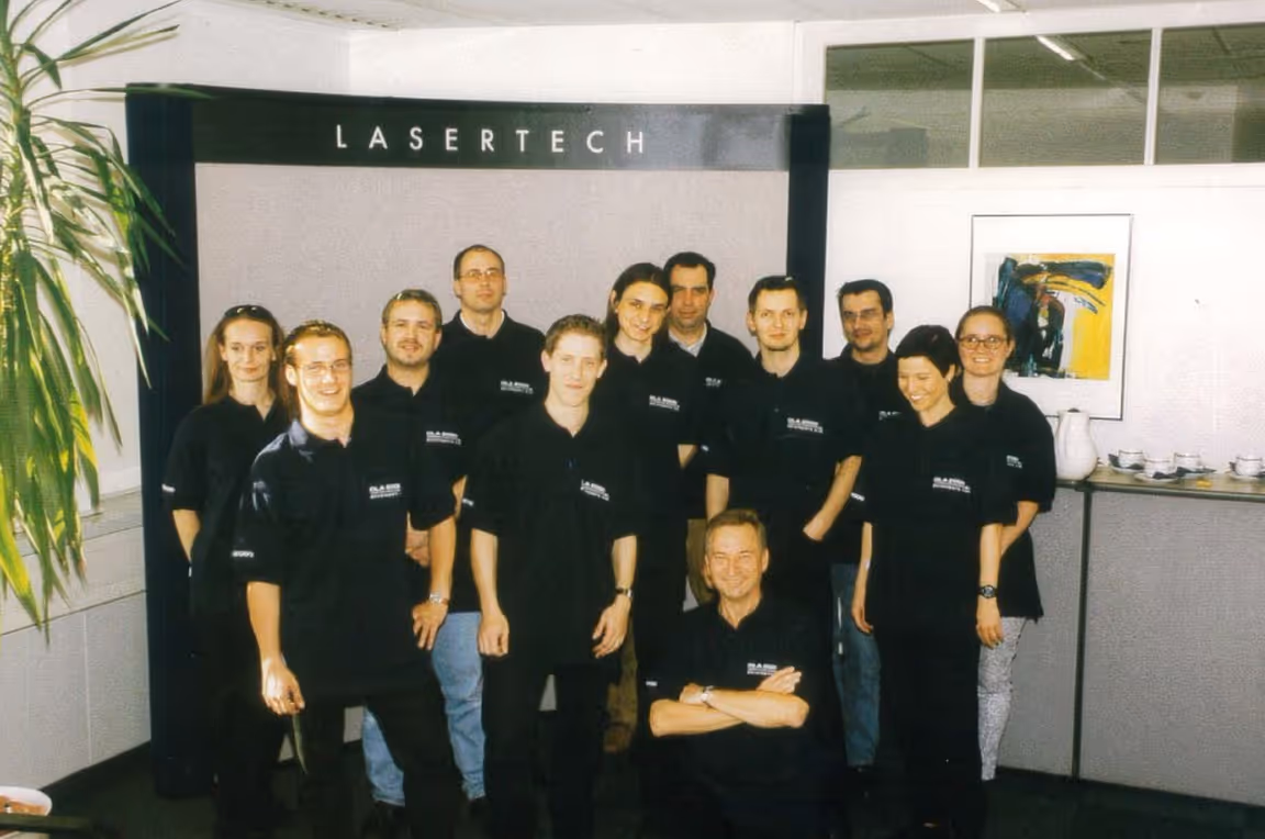 Gruppenfoto von zwölf Personen in schwarzen Firmen-T-Shirts vor einem Schild mit der Aufschrift LASERTECH in einem Büro.