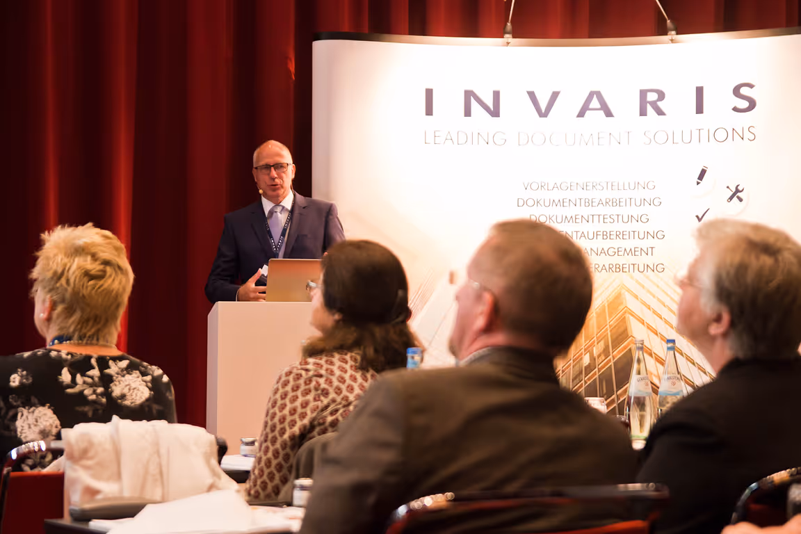 Ein Mann hält einen Vortrag vor einem INVARIS-Banner vor einem Publikum in einem Raum mit roten Vorhängen.
