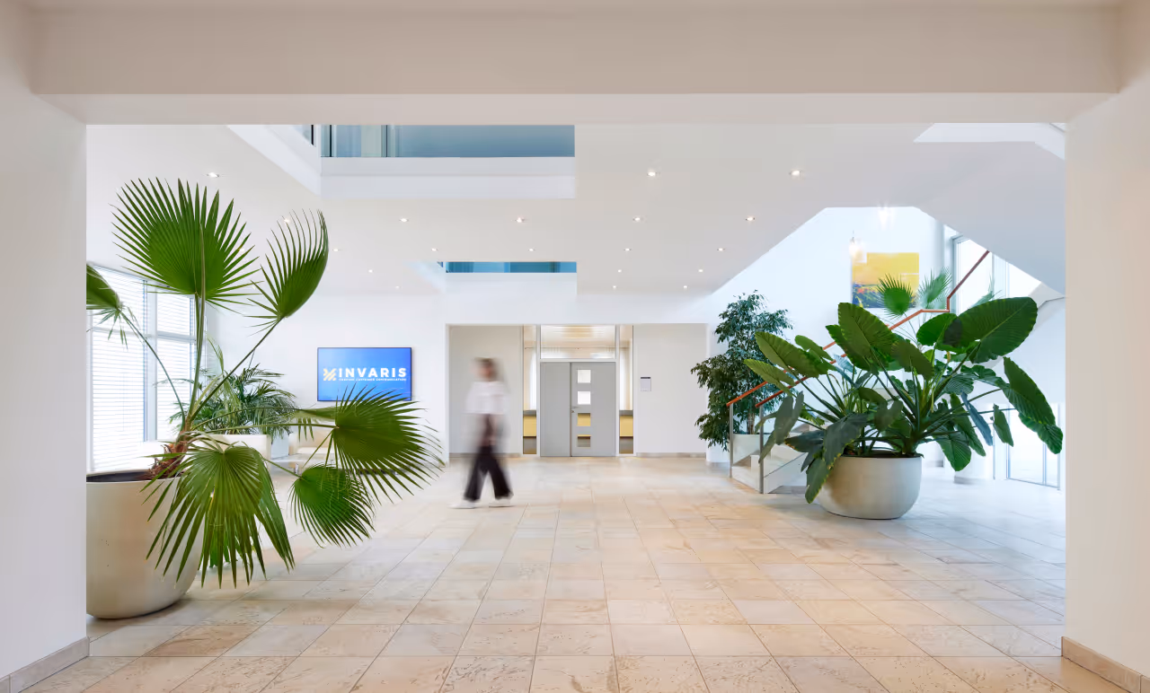 Heller, moderner Büroeingangsbereich mit großen Zimmerpflanzen, Fliesenboden und einer Person in Bewegung neben einem Bildschirm mit dem Logo 'INVARIS'.