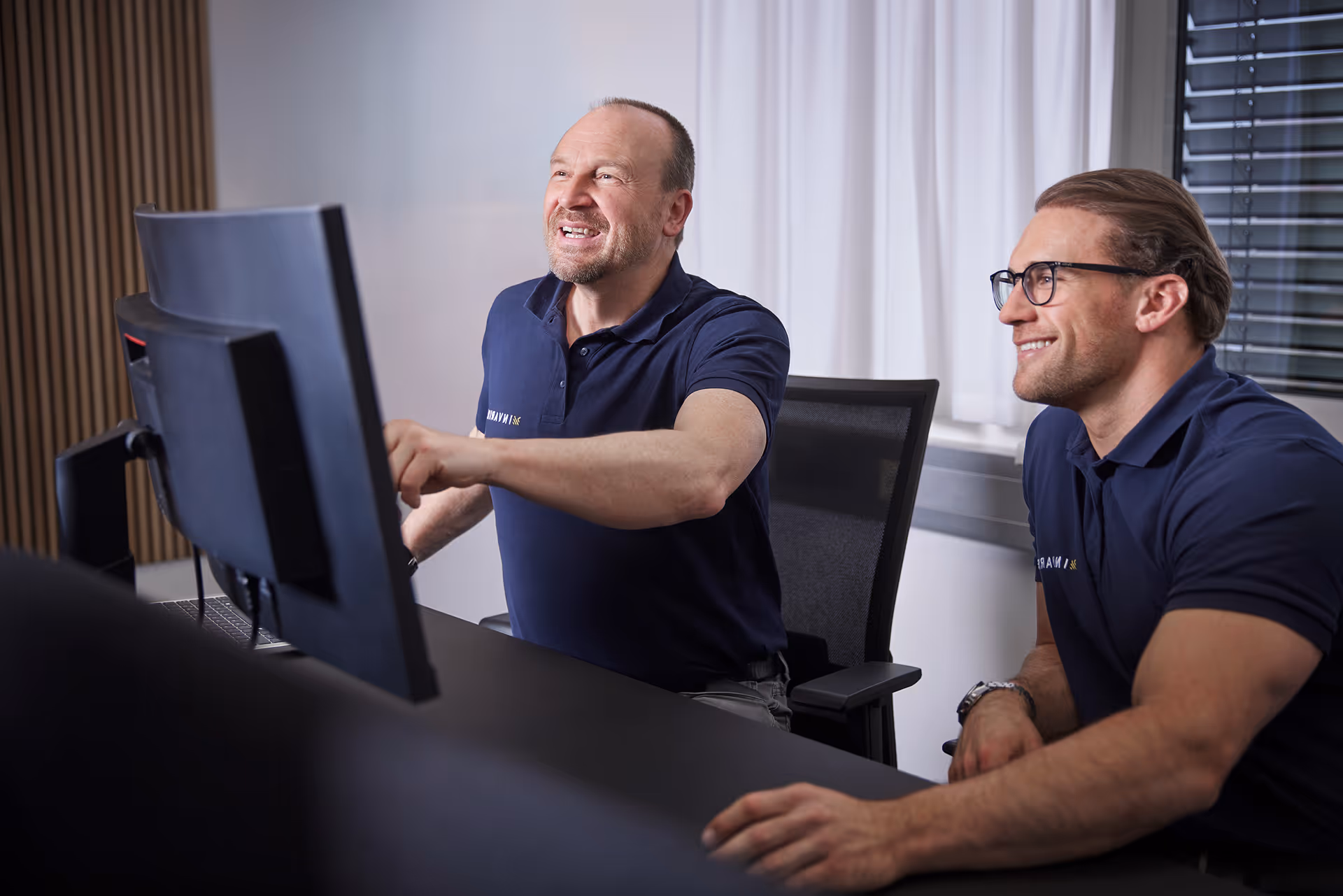 Mann arbeitet konzentriert an einem Laptop in einem Büro mit mehreren Computerbildschirmen im Hintergrund.