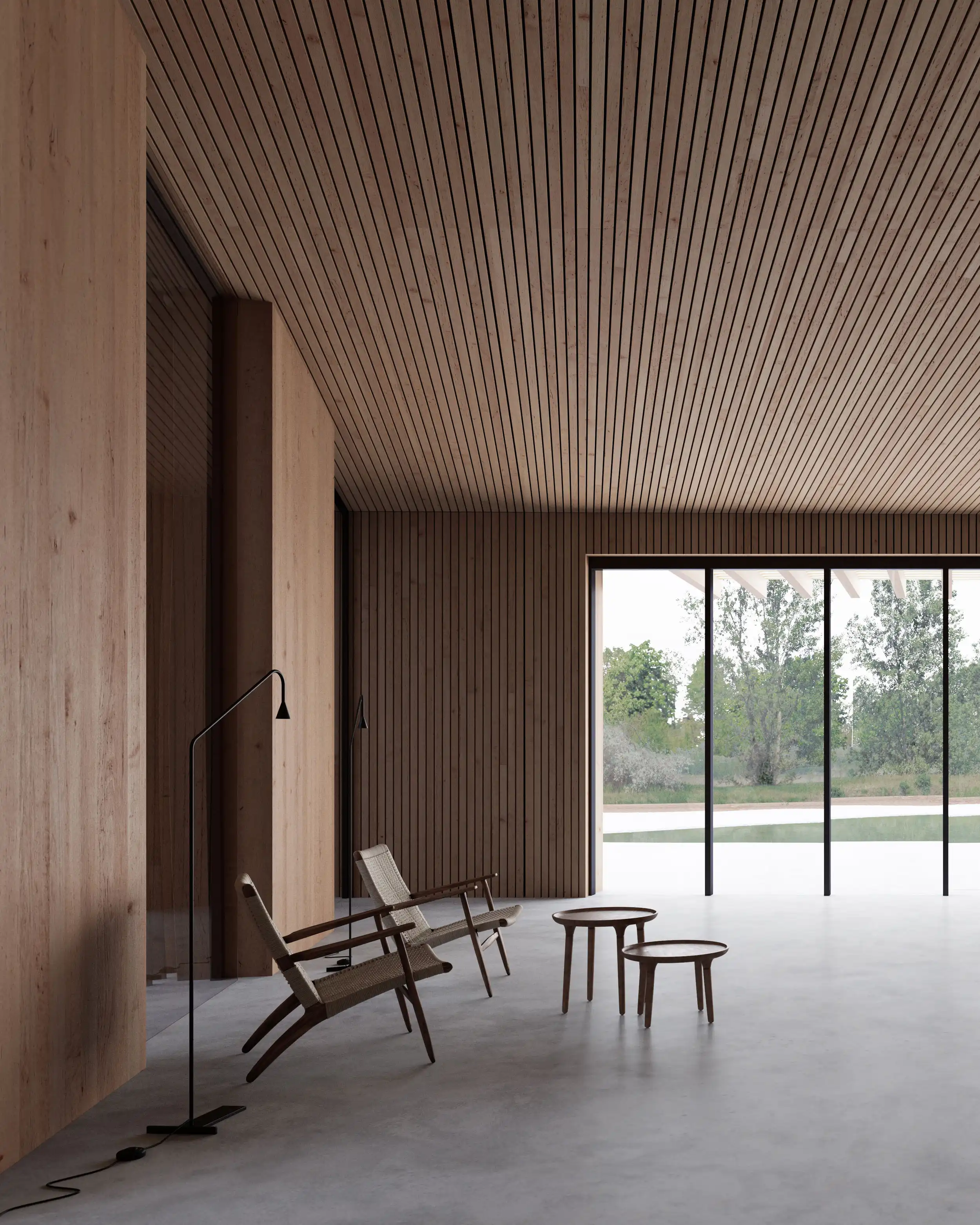 Minimalist interior with wooden slat walls and ceiling, two woven lounge chairs, two round wooden tables, and large floor-to-ceiling glass doors overlooking greenery.