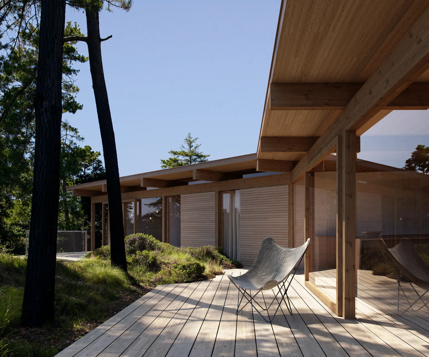 Modern wooden house with large windows and a wooden deck featuring a single gray butterfly chair surrounded by tall trees and shrubs under a clear blue sky.
