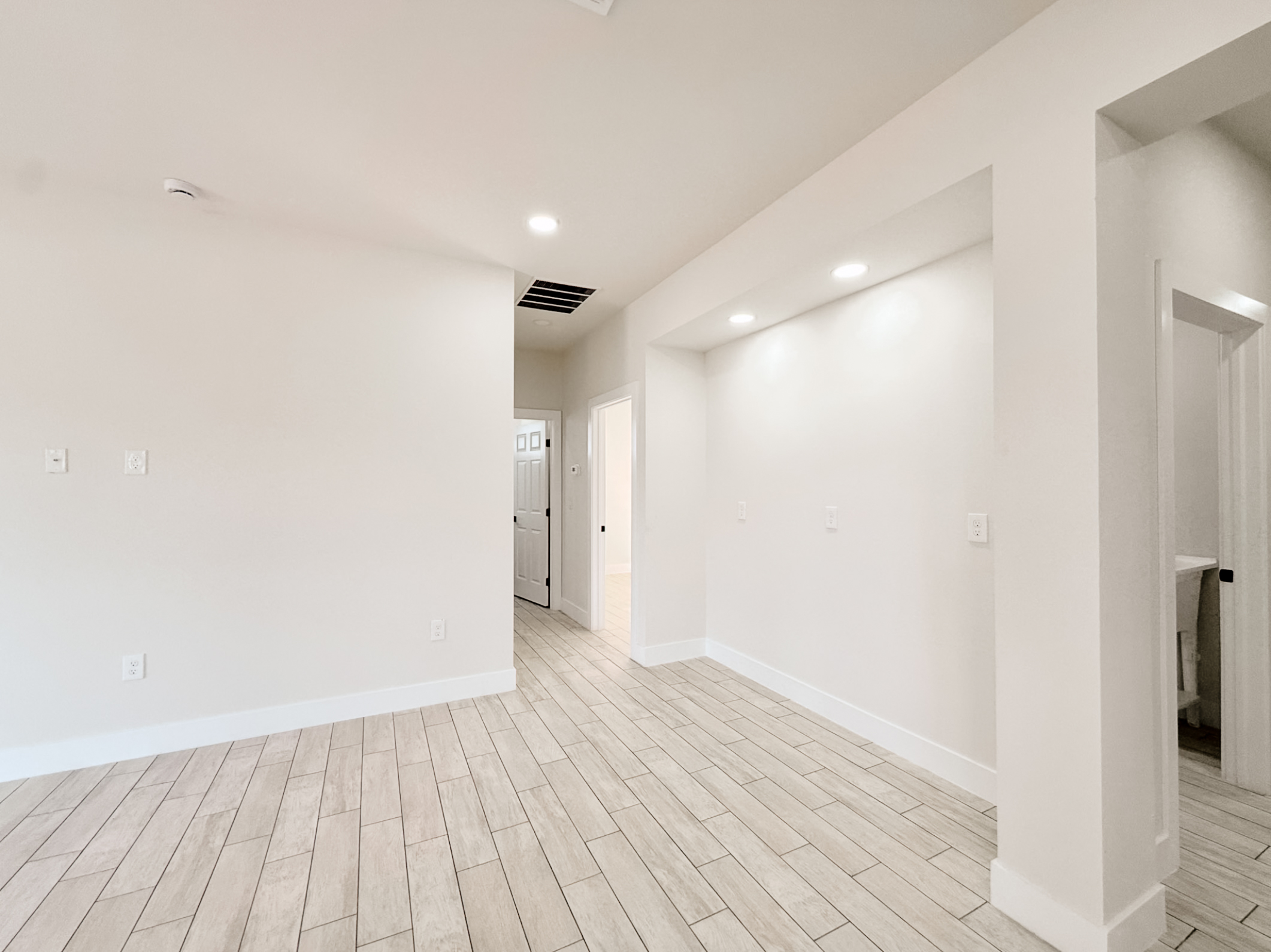 Empty room with light wood tile flooring, white walls, recessed ceiling lights, and open doorways leading to other rooms.