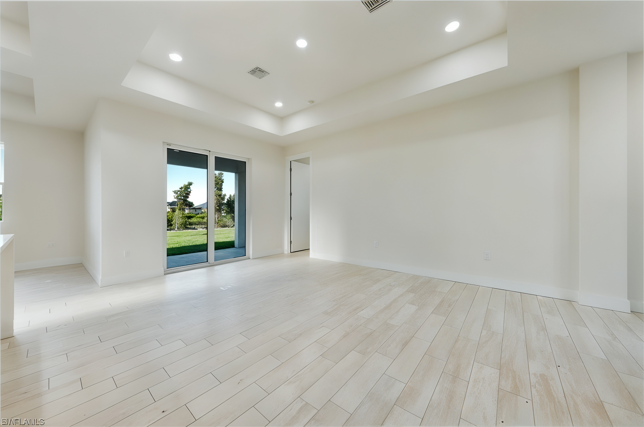 Empty room with light wood-look tile flooring, white walls, recessed ceiling lights, sliding glass doors opening to a green lawn, and an open white door to another room.