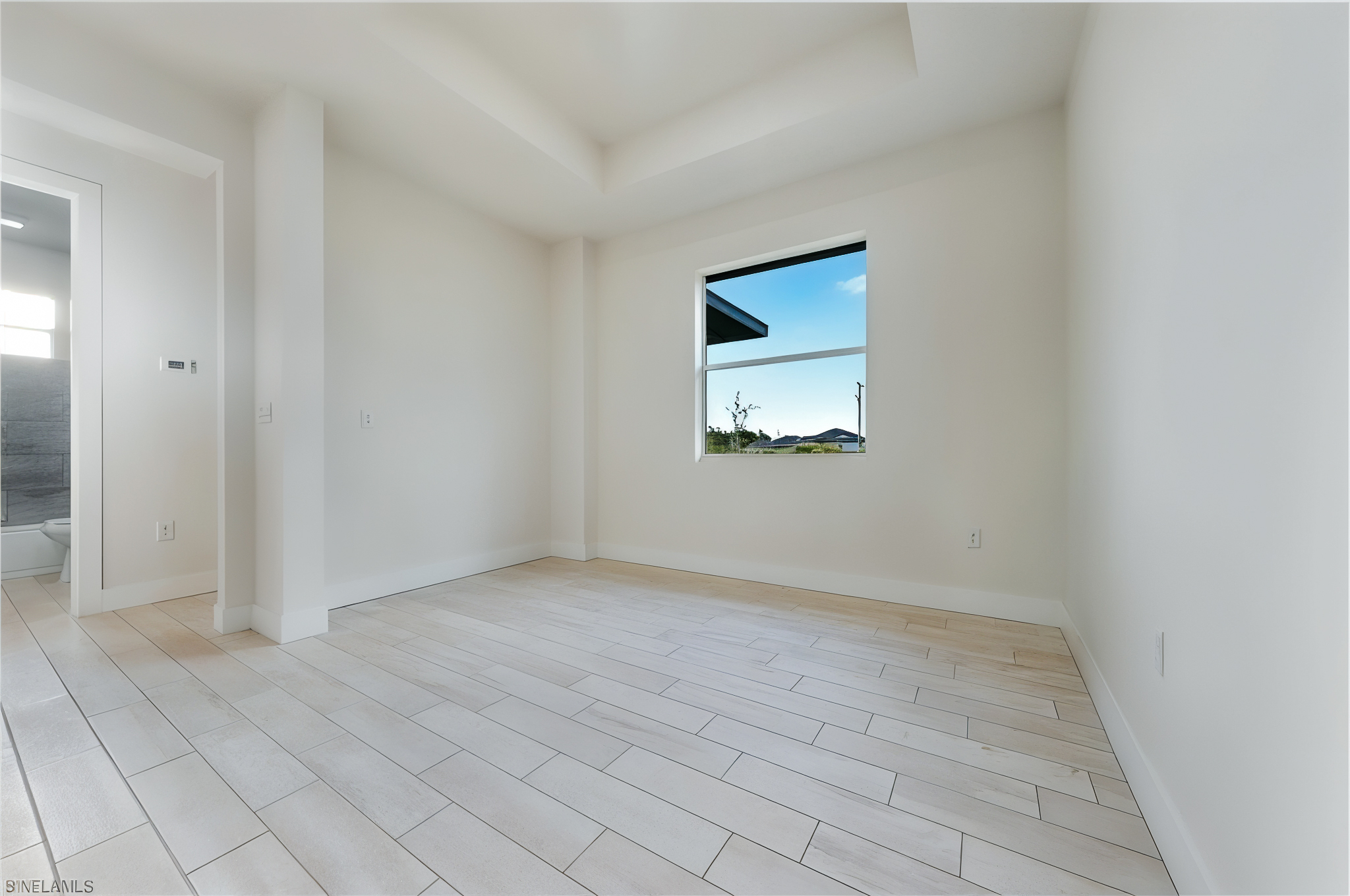 Empty room with light-colored tile flooring, white walls, a window showing an outside view, and an open door leading to a bathroom.