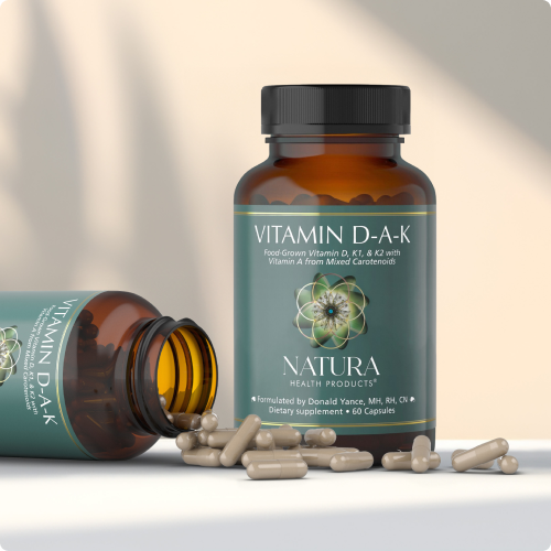 Two brown glass vitamin bottles with teal labels reading Vitamin D-A-K from Natura Health, capsules spill on a pale surface.