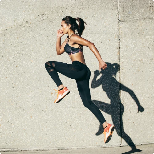 Athletic woman in a sports bra and black leggings jumps mid-air beside a concrete wall, orange sneakers and ponytail shadow.