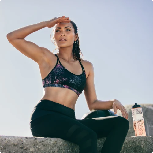 Sunlit athletic woman sits on a ledge, shading her eyes with hand, wearing a black patterned sports bra and leggings; a water bottle rests nearby.