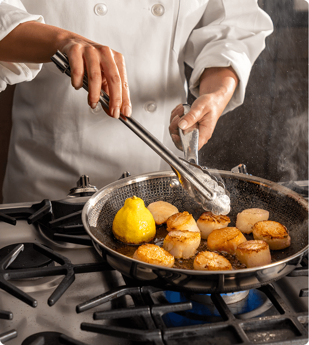 Chef using tongs to cook seared scallops and a halved lemon in a frying pan over a gas stove.