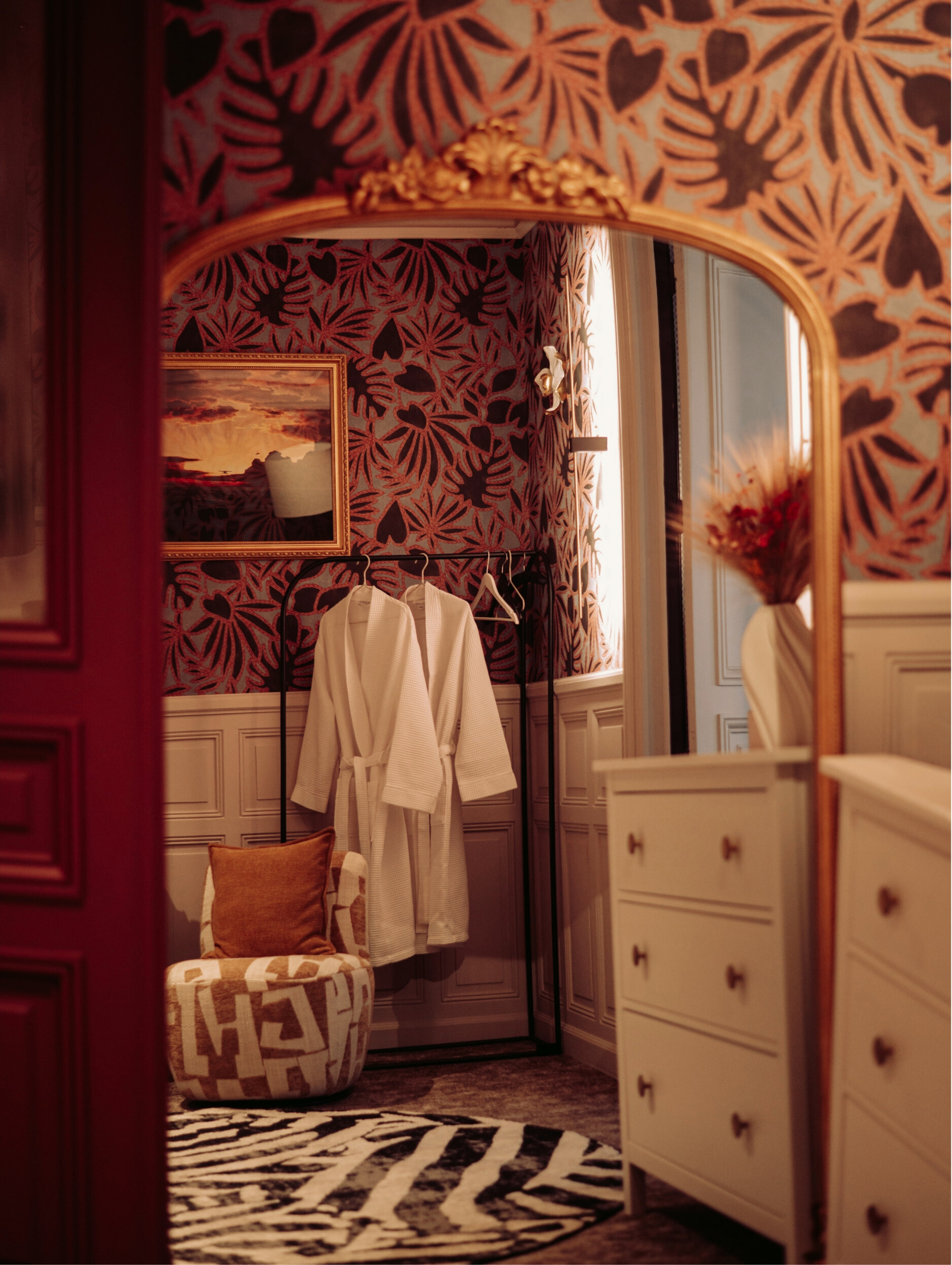 Chambre avec des peignoirs blancs suspendus, un fauteuil imprimé beige avec coussin, un tapis zèbre, papier peint floral et une commode blanche reflétée dans un miroir doré.