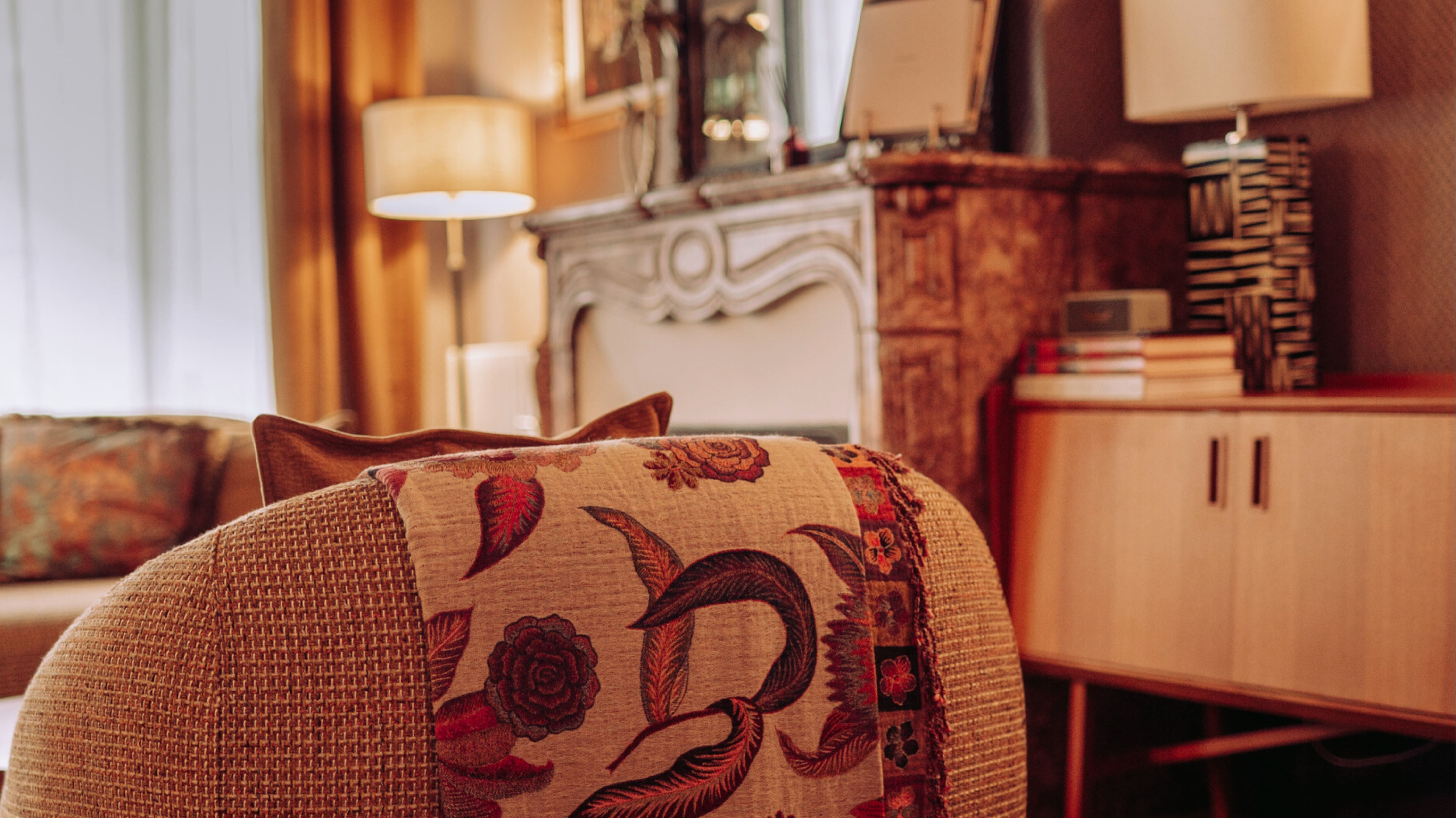 Fauteuil avec couverture à motifs floraux en premier plan, dans un salon chaleureux avec lampe, cheminée décorative et buffet en bois à l'arrière-plan.