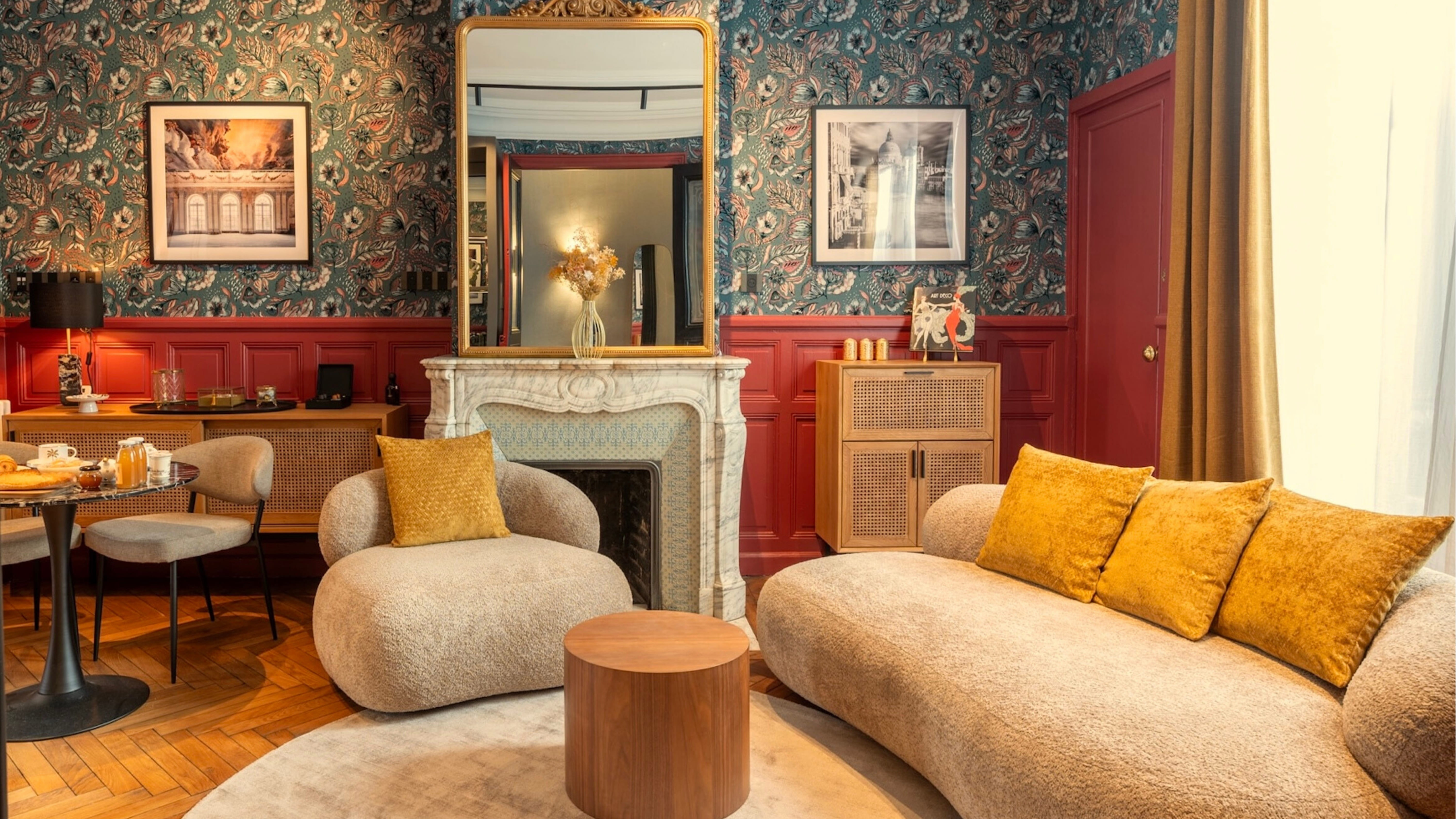 Salon avec canapé beige et coussins jaunes, fauteuil assorti, table basse ronde en bois, cheminée en marbre, et murs décorés de papier peint floral et cadres.