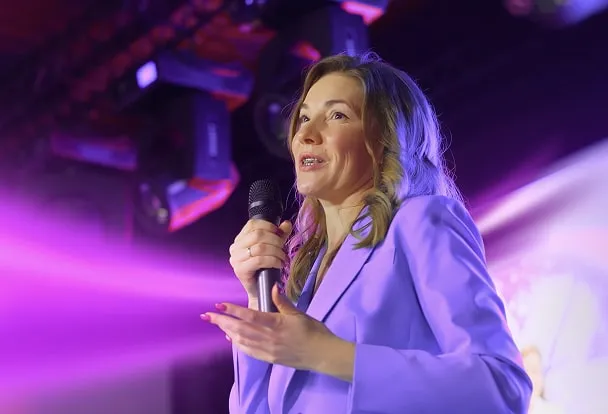 Woman in a lavender blazer speaking into a microphone on stage with purple lighting.