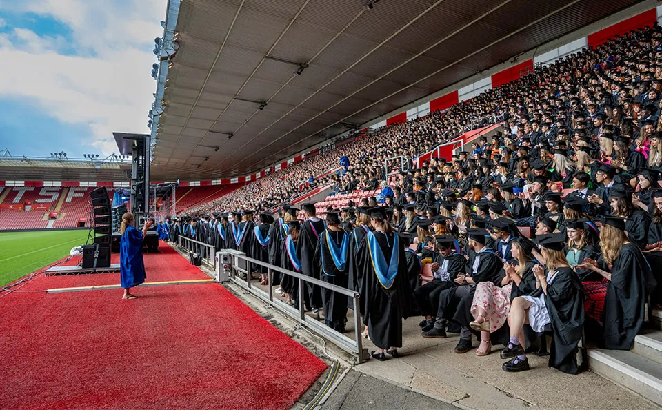 Graduates in caps and gowns lined up at a stadium event with many spectators seated in the stands.