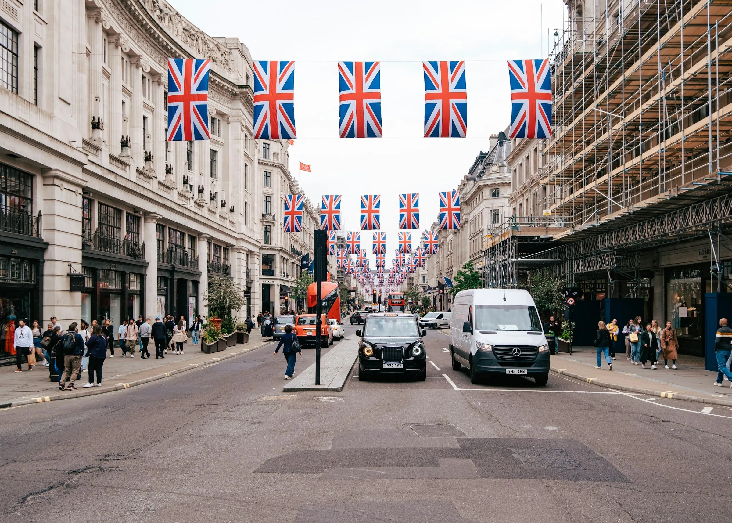 Busy street decorated with hanging Union Jack flags, lined with historic buildings, pedestrians, and vehicles including a black taxi and white van.