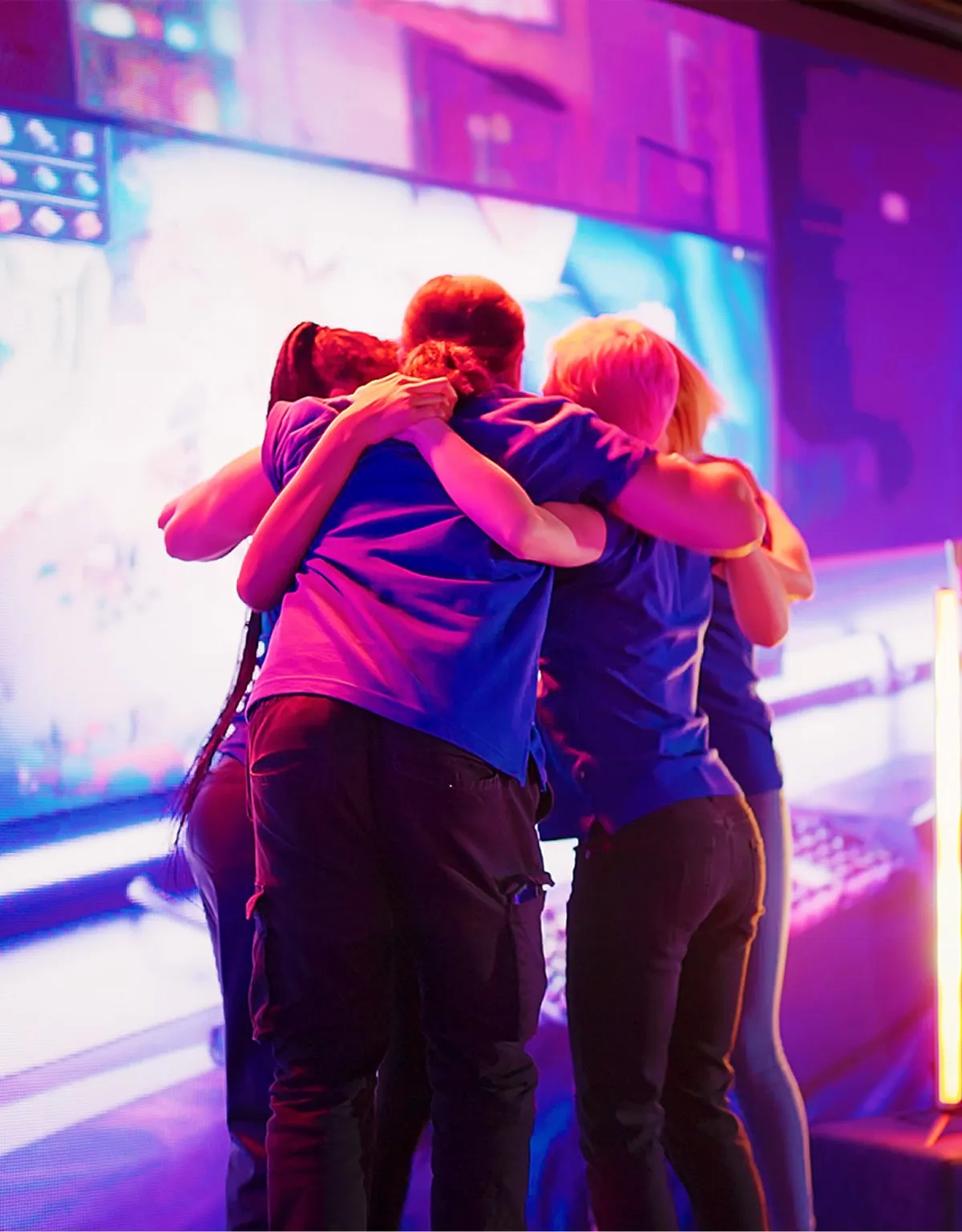 Group of four people in blue shirts hugging each other in front of a large screen with purple and blue lighting.